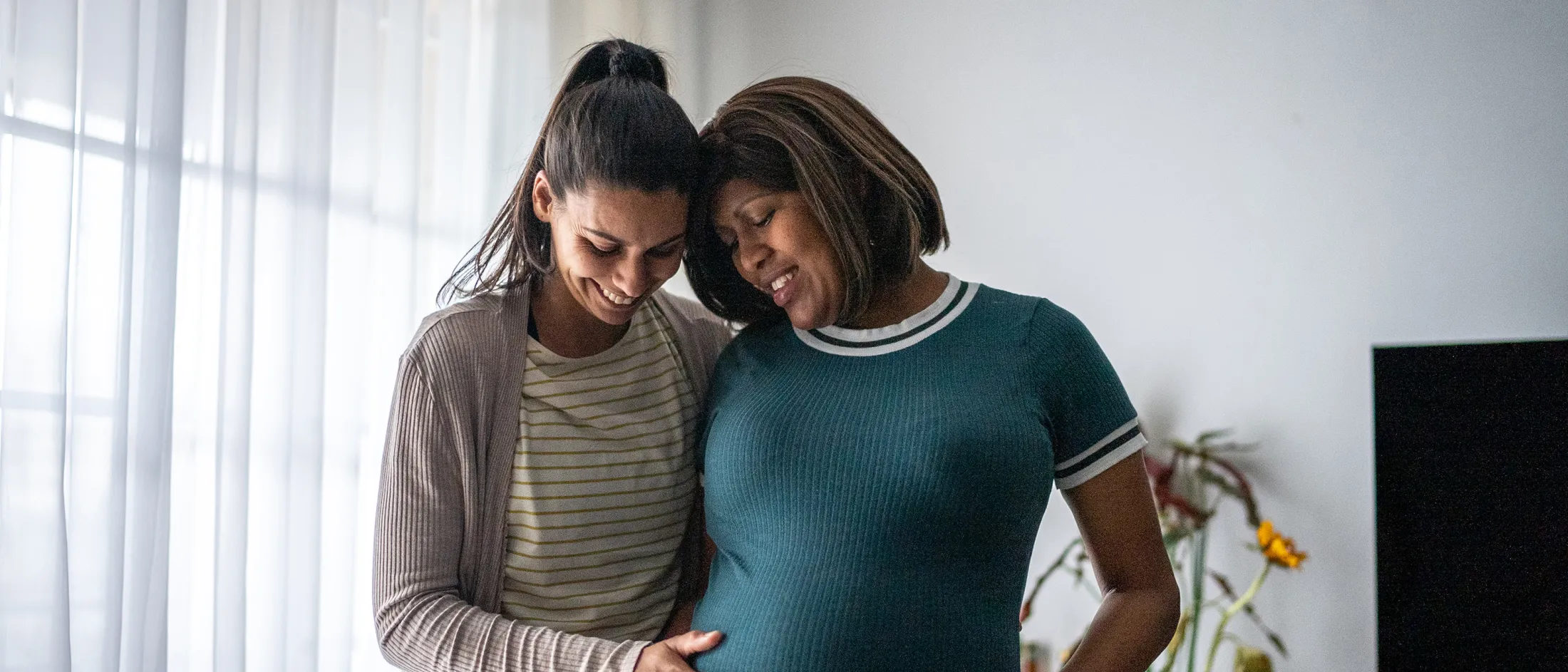 Two women smiling and embracing while touching a pregnant belly in a warmly lit home environment.