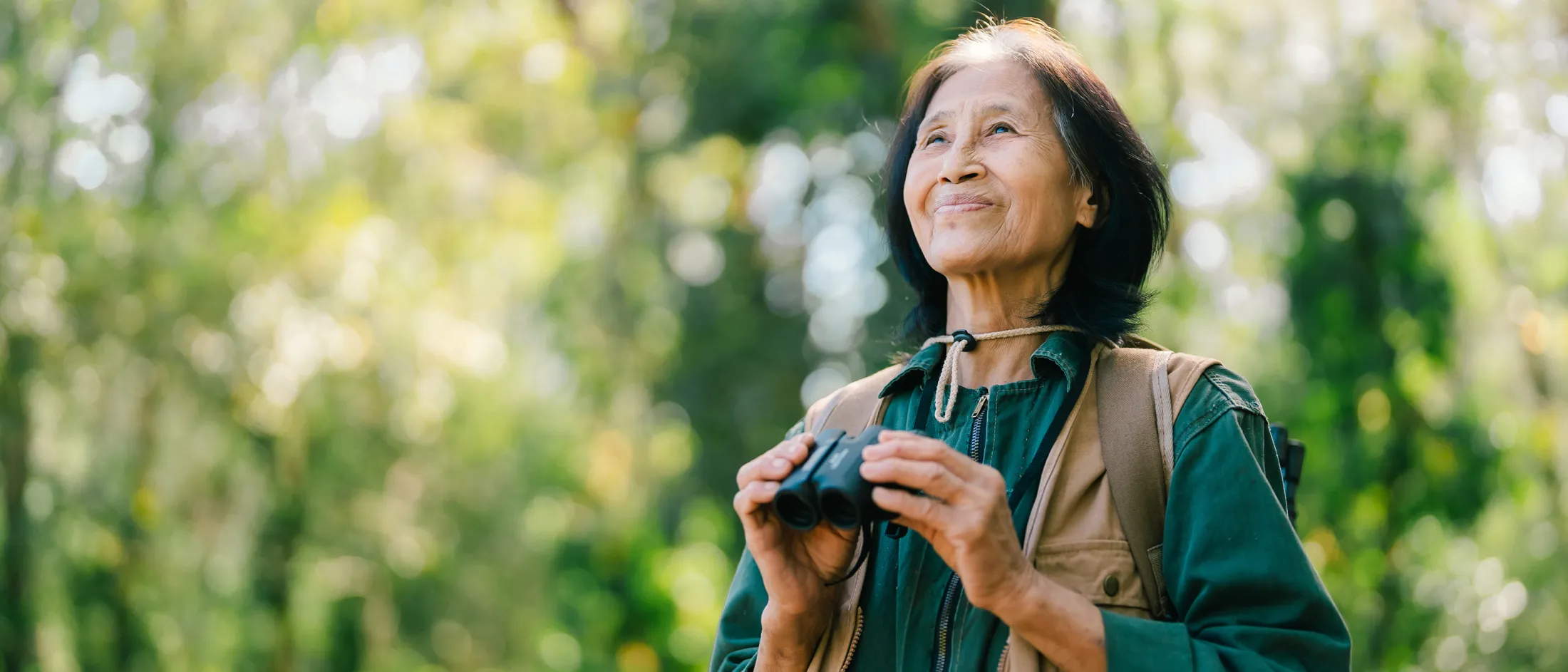 Older woman in outdoor clothing holding binoculars and gazing upward while birdwatching in a lush green forest.