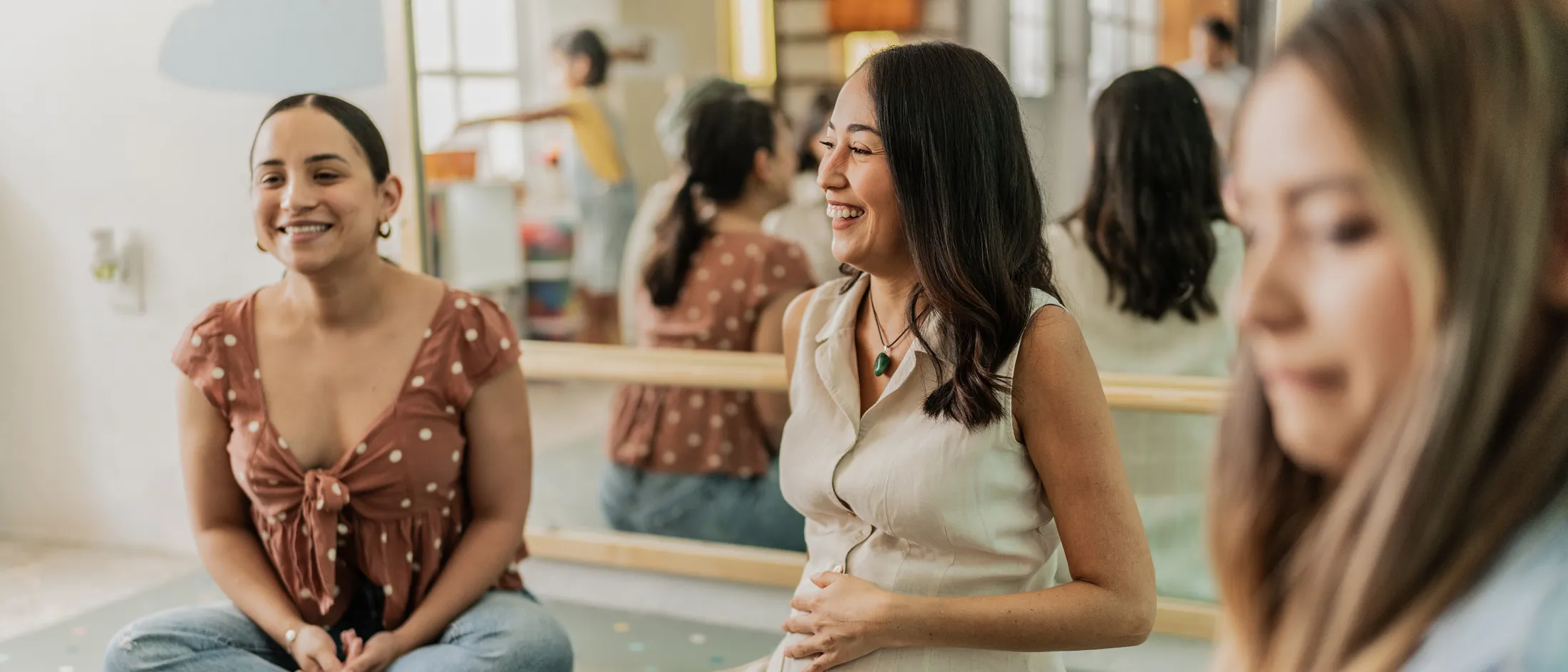 Pregnant woman smiling during a group session with other women in a brightly lit room with large mirrors.