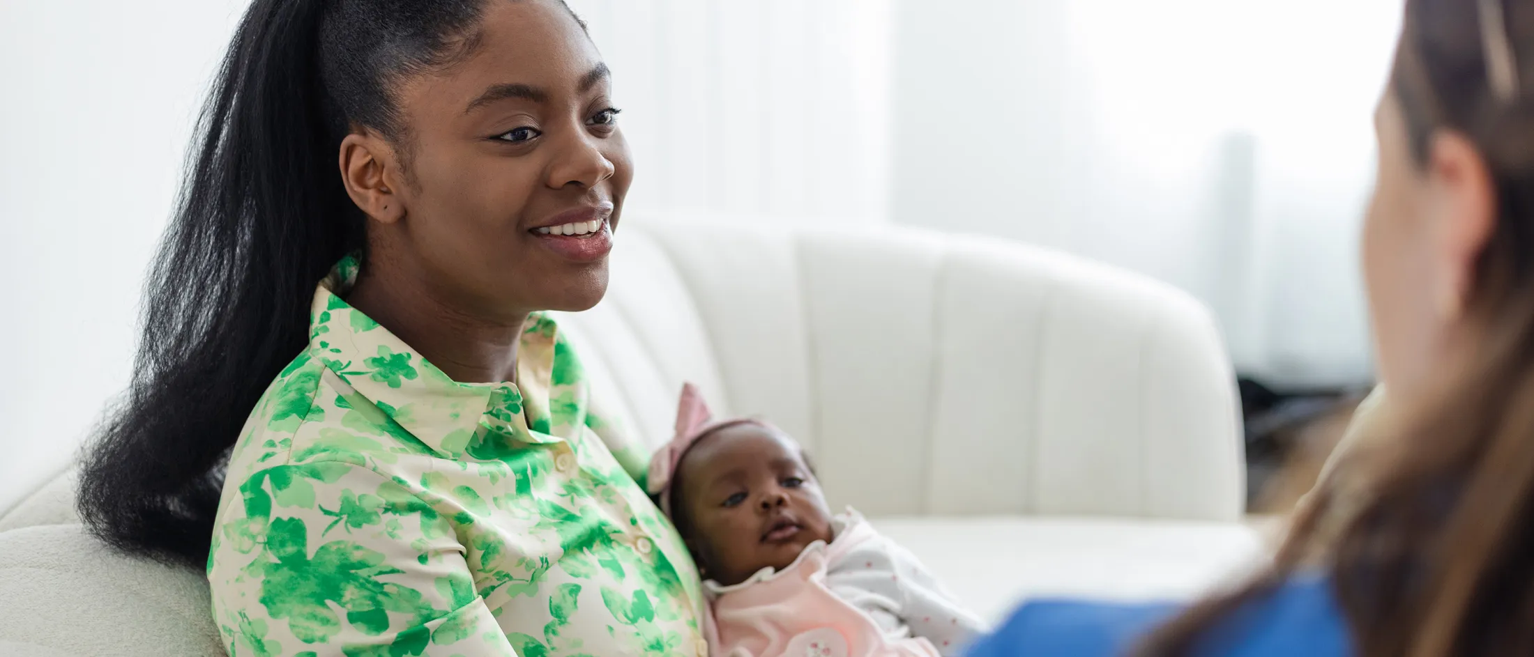 Woman with long hair in a green floral blouse holding a baby while talking to another adult in a light-filled room.