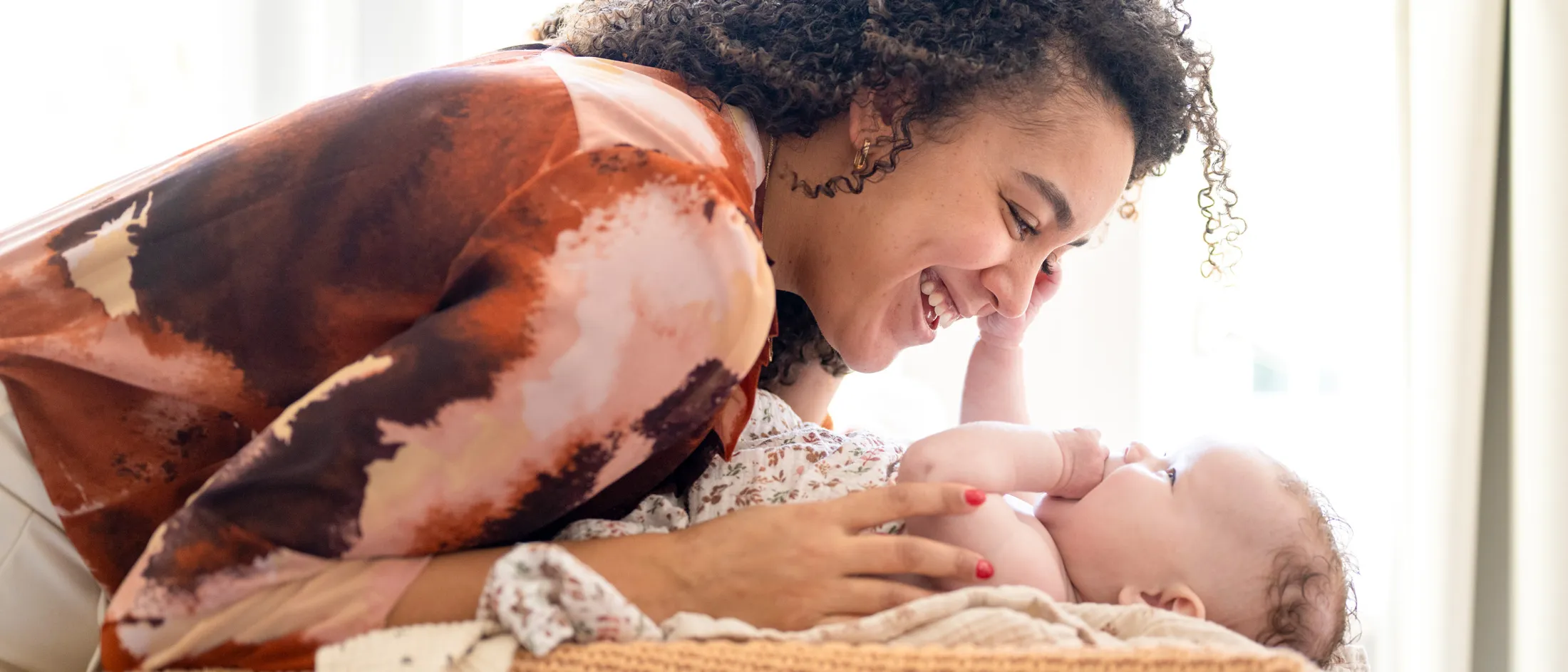 A smiling woman with curly hair leans over a baby, engaging in joyful eye contact while lying together on a bed or couch.