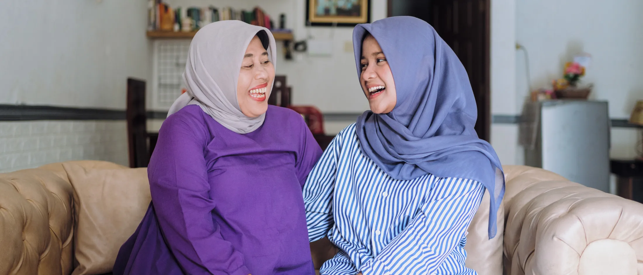 Two women wearing hijabs laughing and enjoying a joyful moment together on a living room couch.