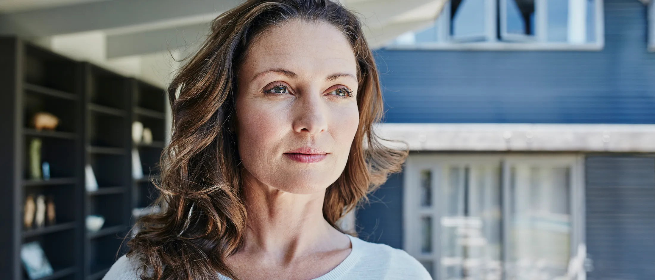 A middle-aged woman with wavy brown hair stands indoors, gazing thoughtfully out a window with soft natural light highlighting her face.