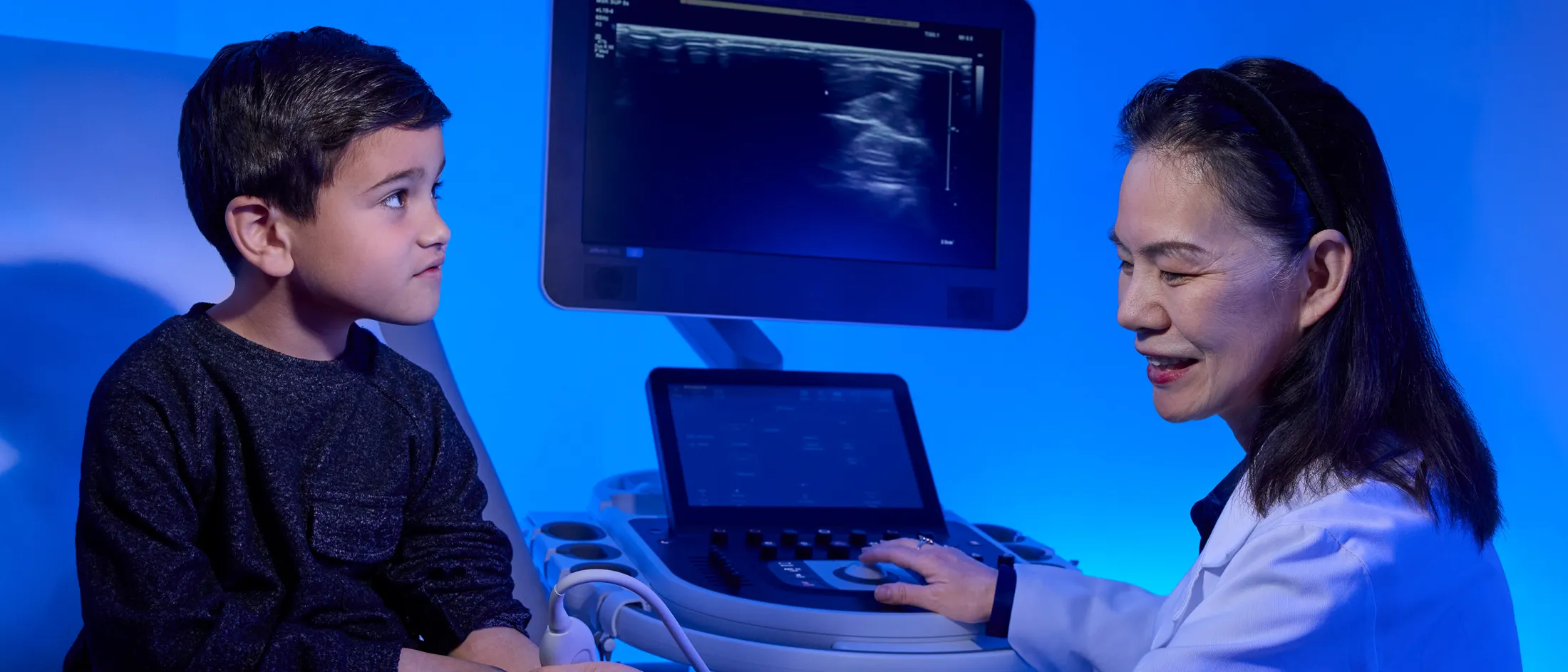 Female doctor performing an ultrasound on a young boy’s wrist in a brightly lit exam room.