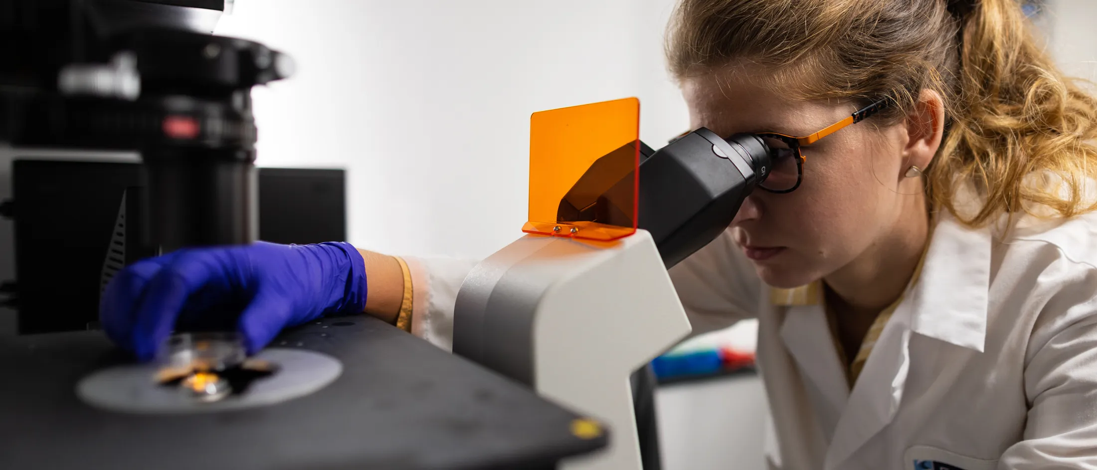 Female researcher in a lab coat using a high-powered microscope, adjusting a sample with a gloved hand.
