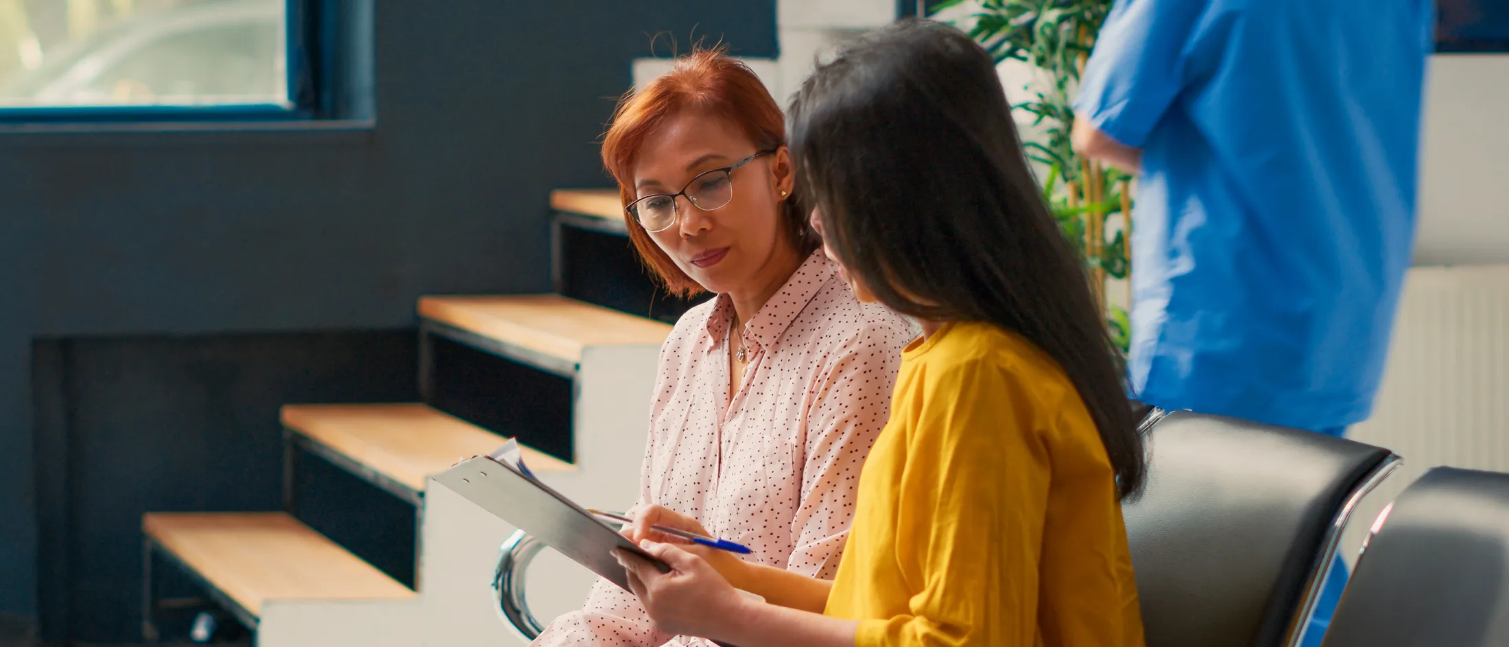 Two women discussing a document while seated in a hospital waiting area, with medical staff in the background