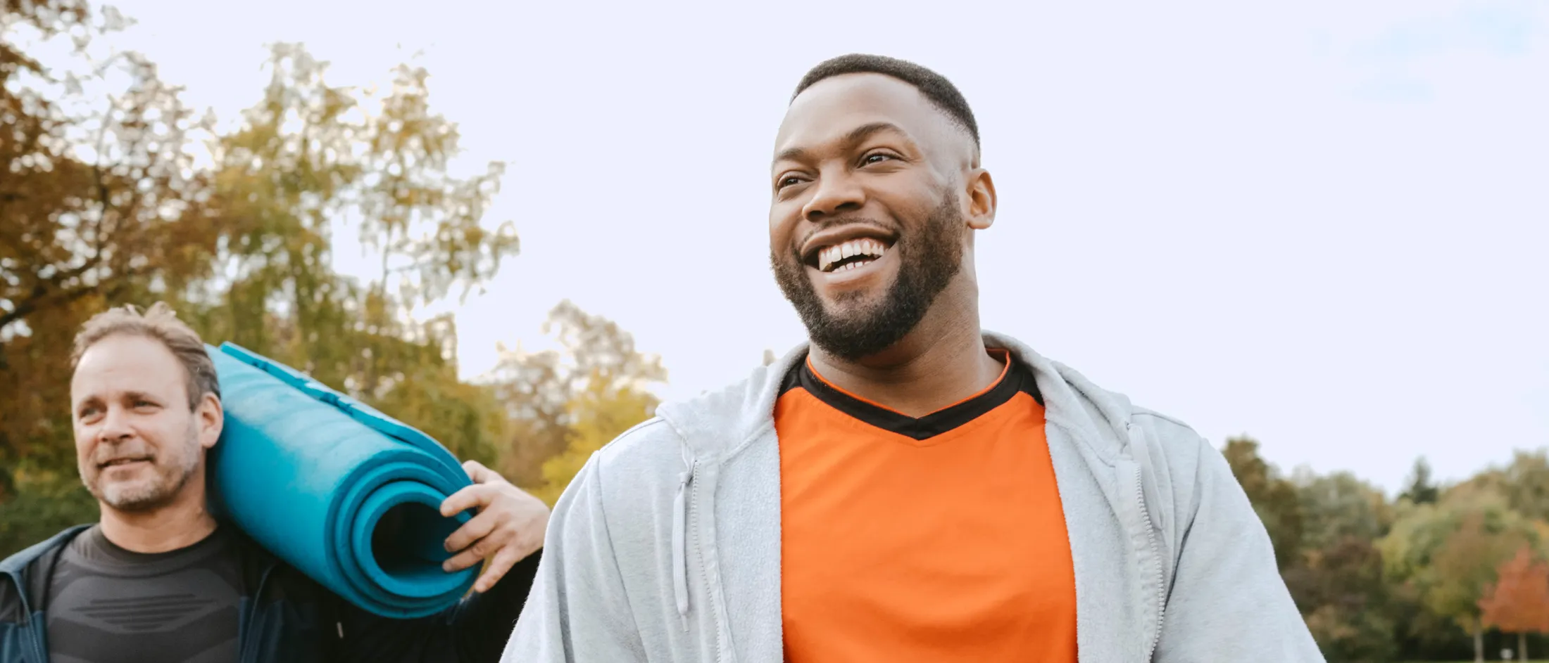 Two men outdoors, one carrying a yoga mat and the other smiling in an orange athletic shirt.