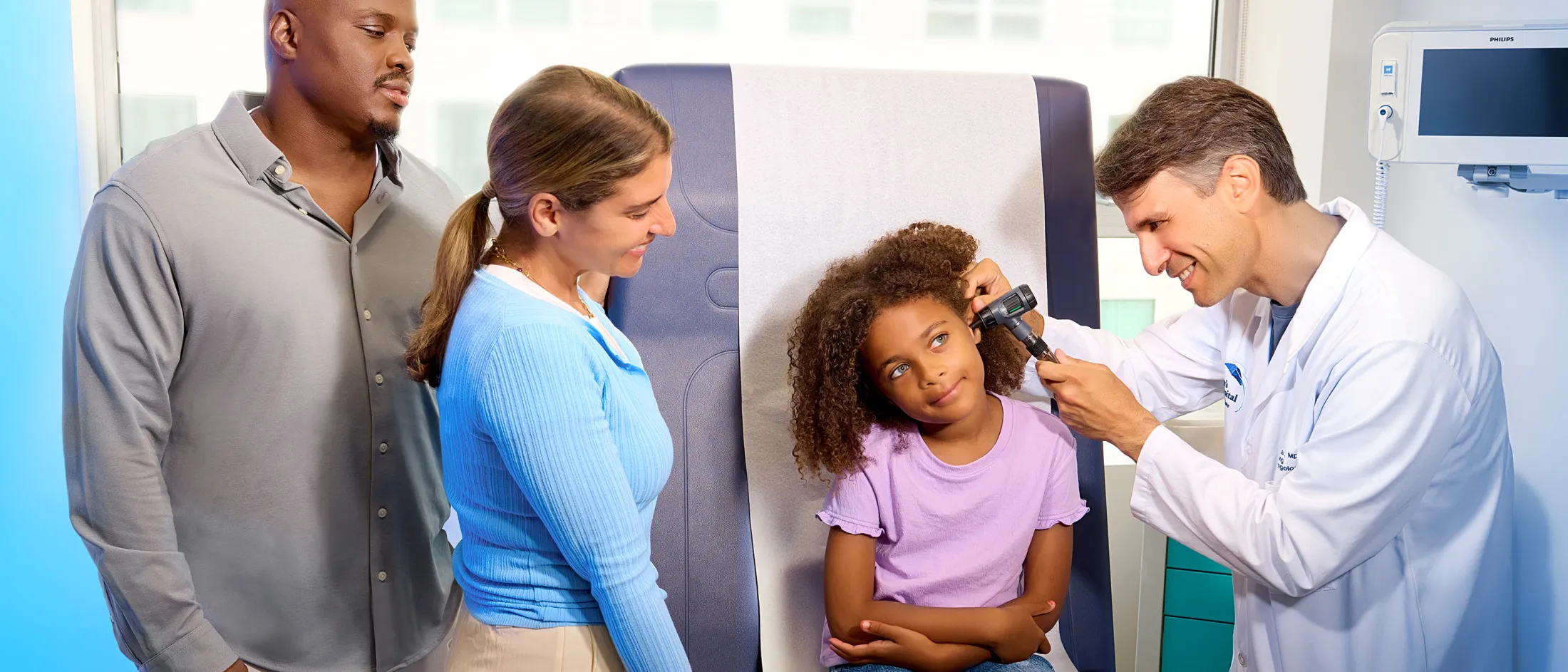 Pediatrician examining a young girl’s ear with an otoscope while her parents stand nearby, smiling and engaged in a bright clinic exam room.
