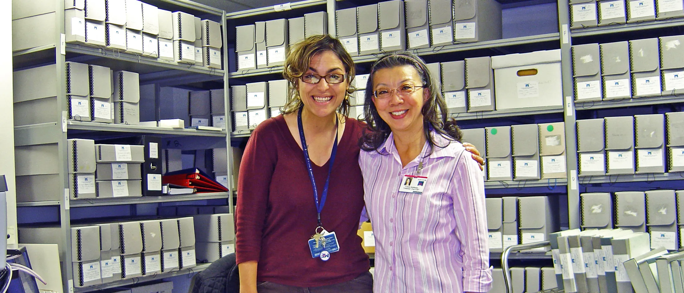 Two women smiling and standing together in an archive room filled with labeled boxes and shelves