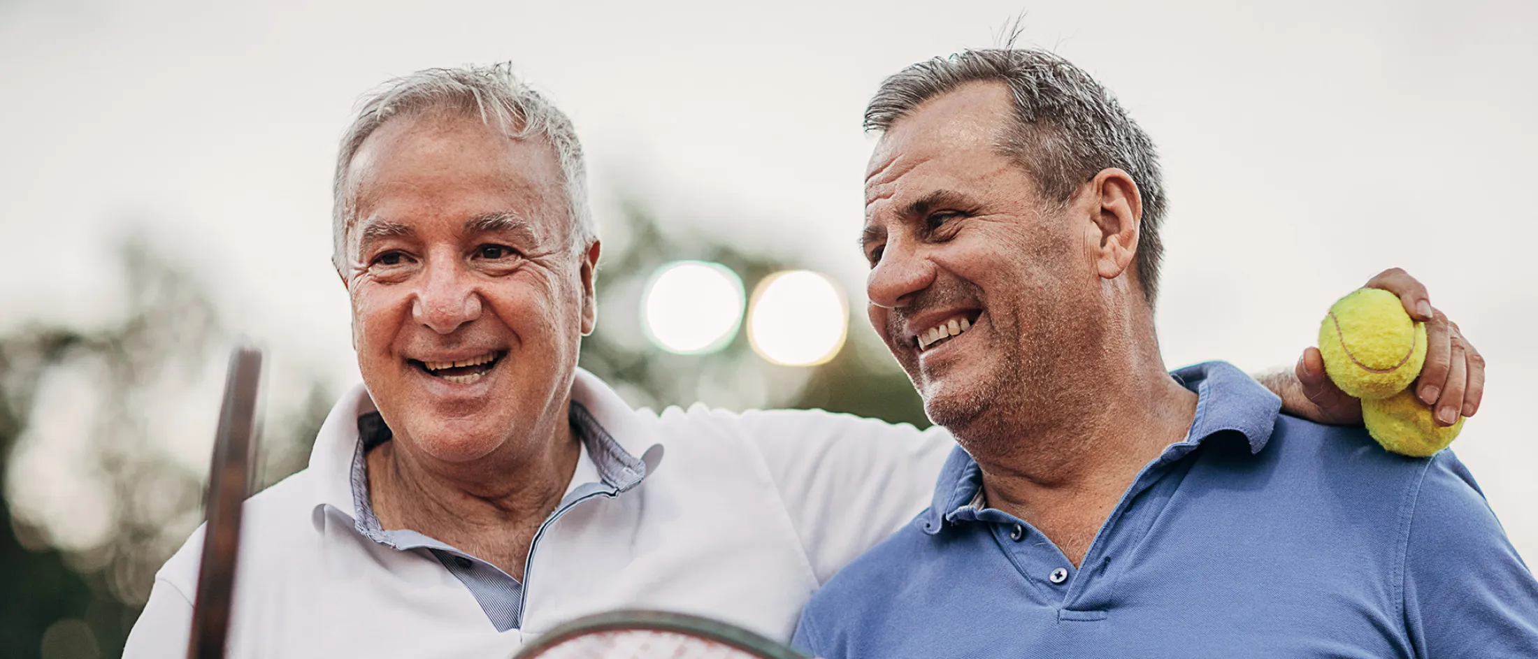 Two smiling men holding tennis gear stand side by side on a court