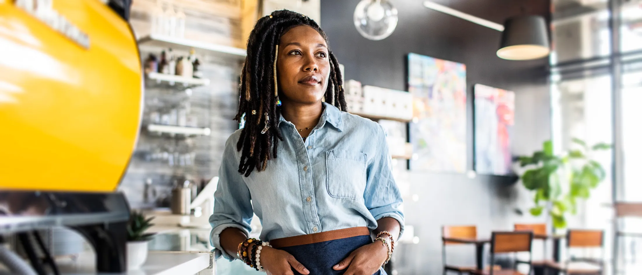 A confident woman in a denim shirt and apron stands behind the counter of a stylish café, looking thoughtful and composed as sunlight filters through large windows.