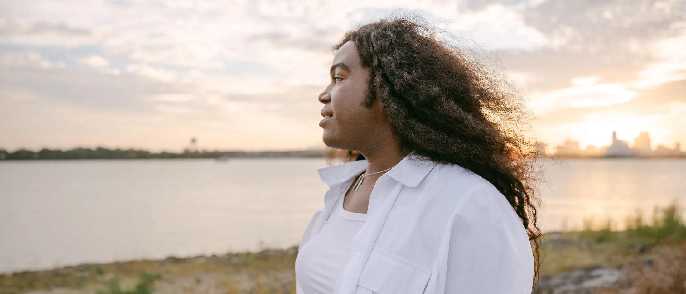 A transgender woman in a white shirt gazes thoughtfully across the water at sunset.