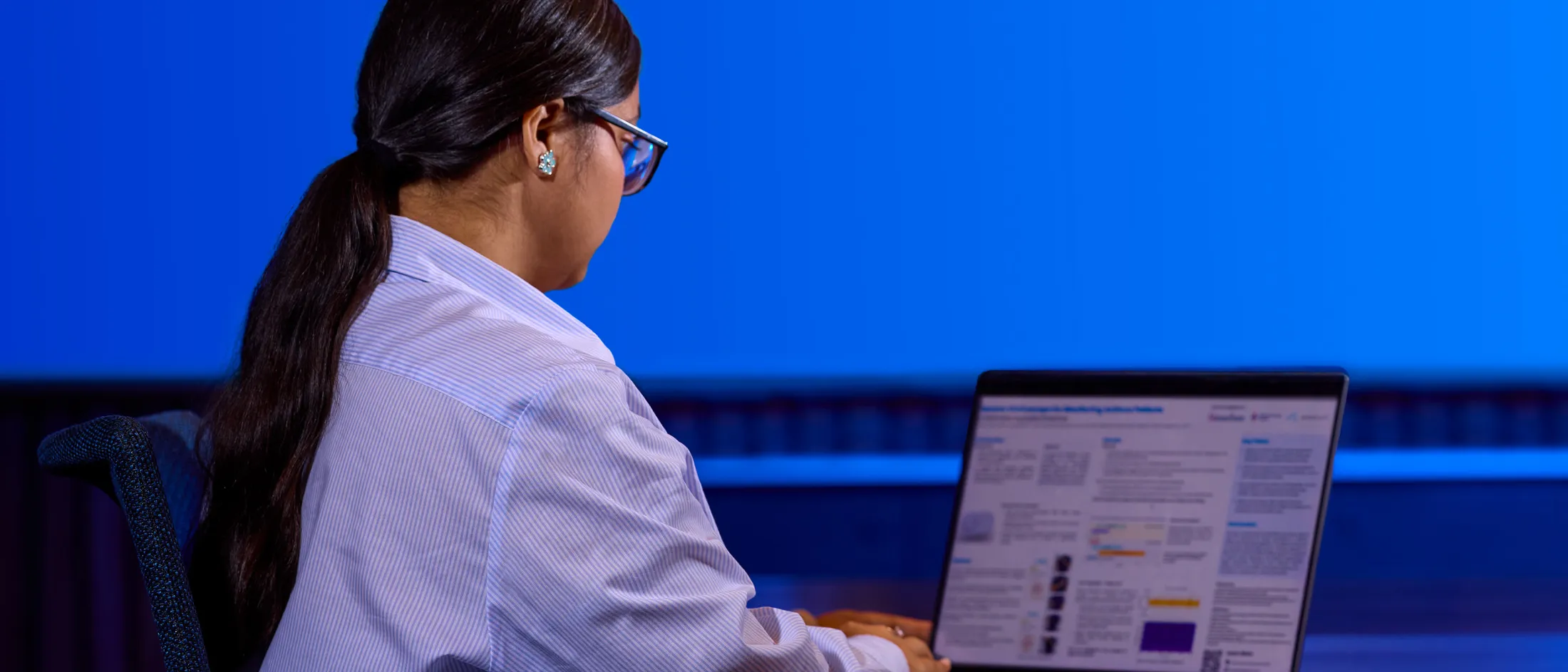 Woman with long dark hair reviews academic research on a laptop in a quiet study space with blue lighting.