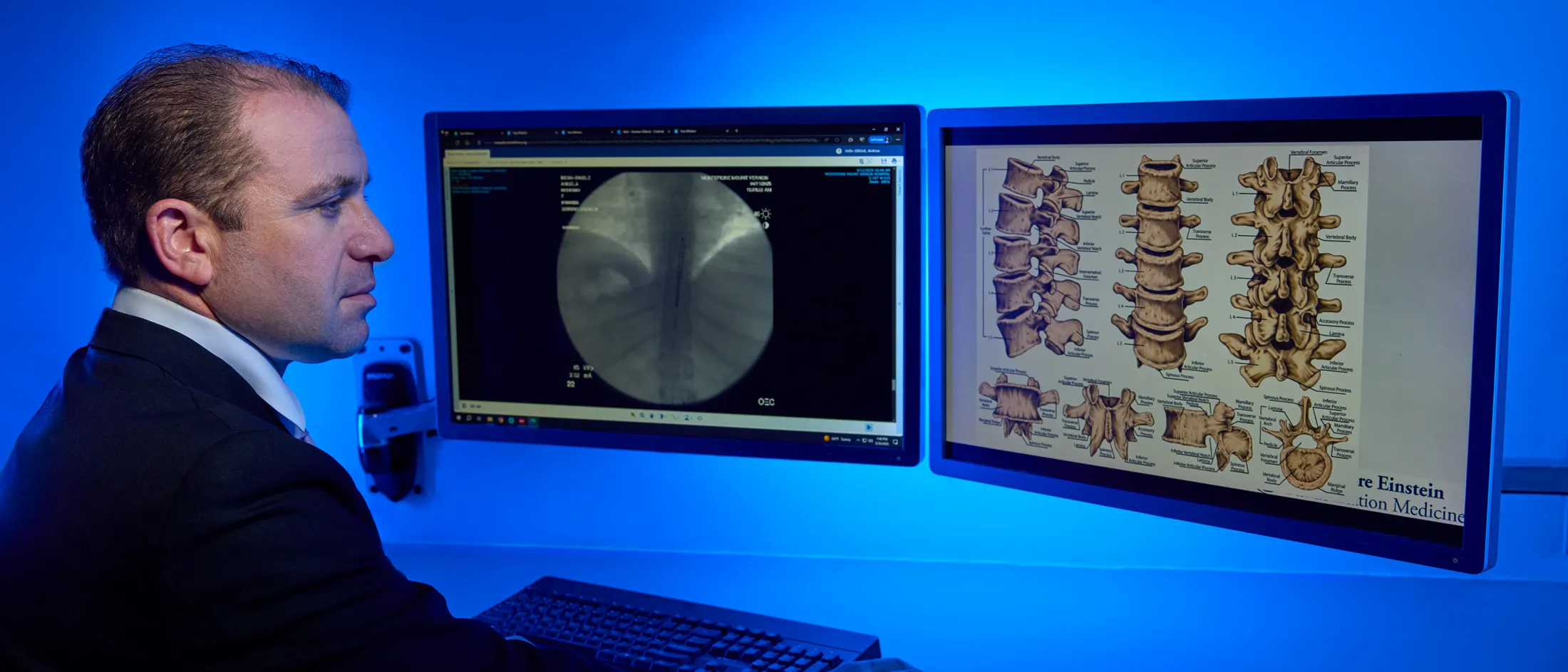 Physician studies spine anatomy illustrations and radiographic images displayed on dual monitors.