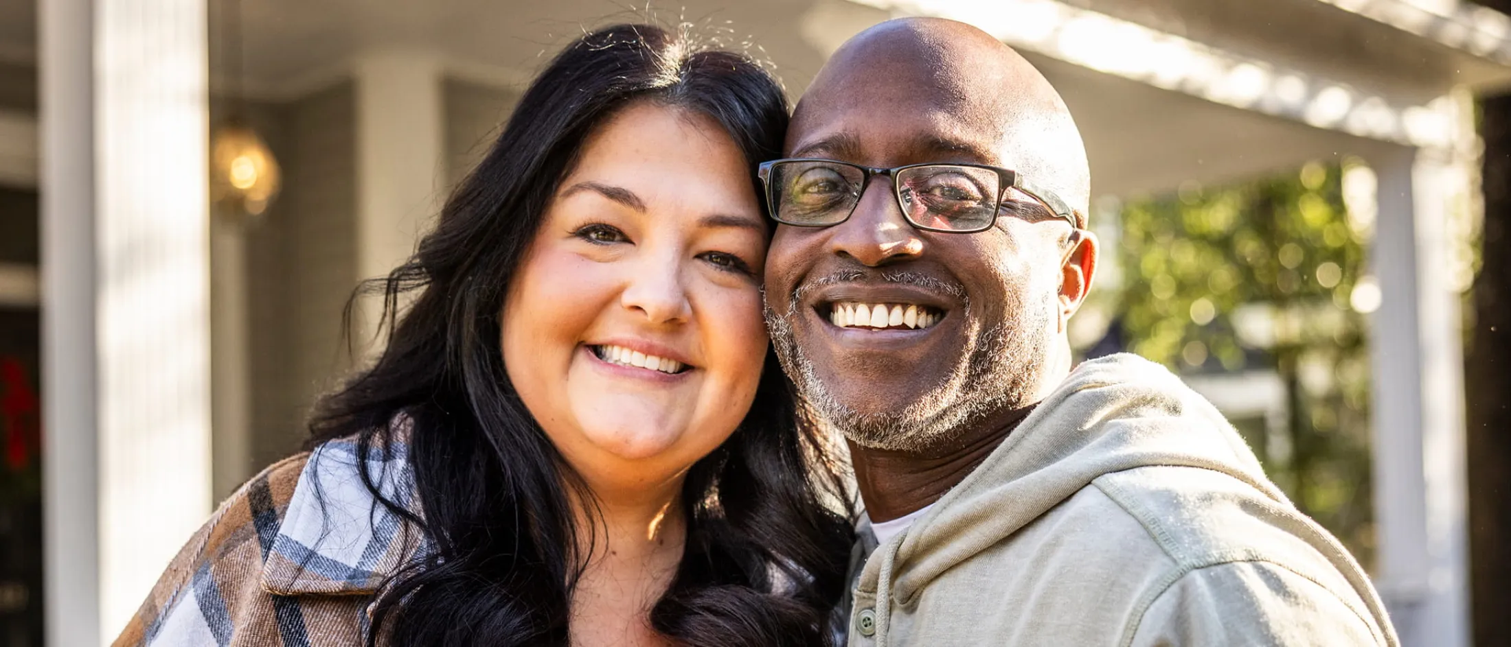 Man wearing glasses, smiling next to a woman as they stand in front of a house