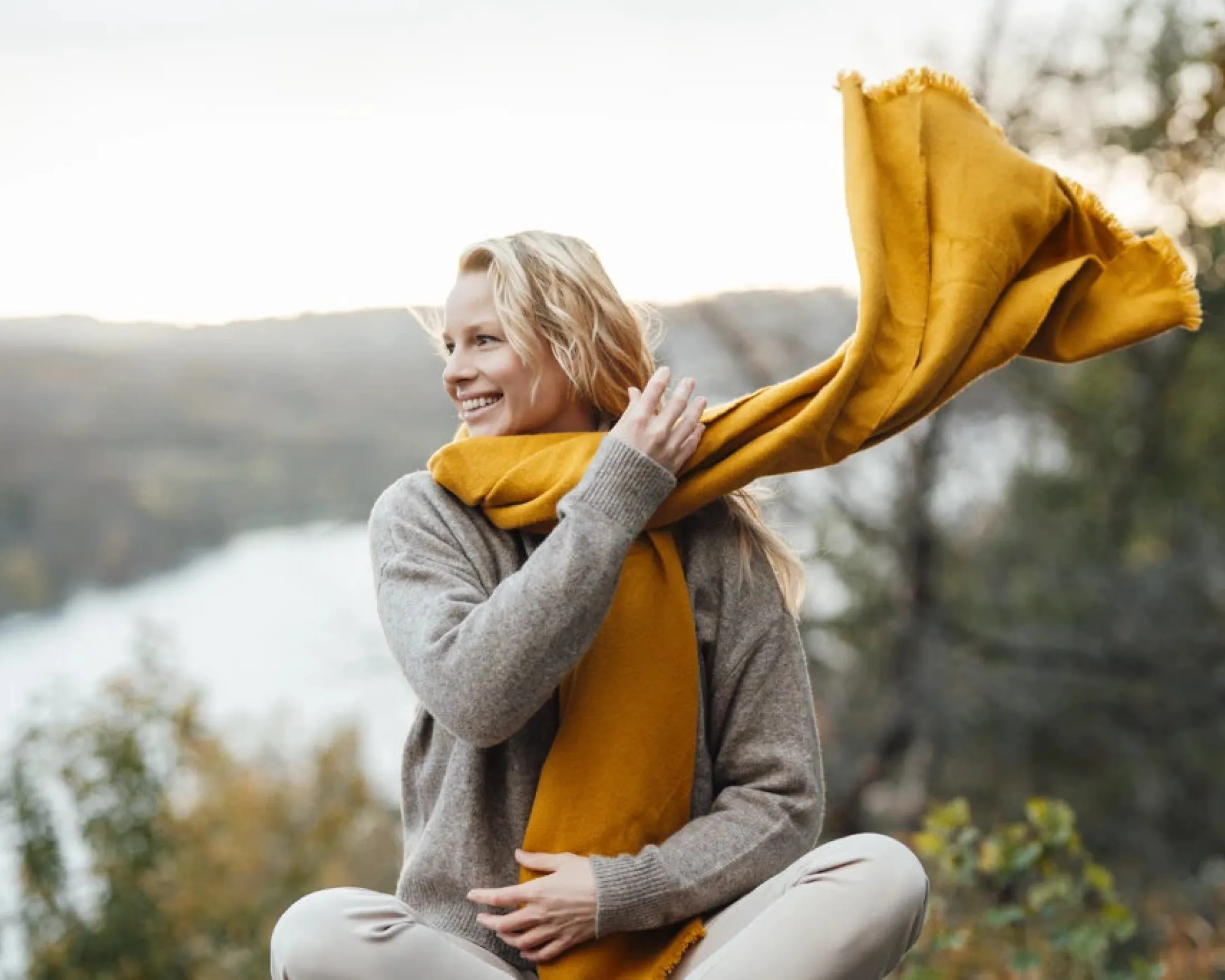 A blonde woman sitting in nature. She is holding a yellow scarf that is blowing in the wind.