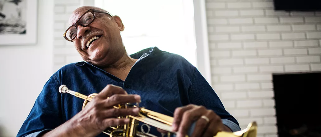 A middle-aged black man in tortoiseshell glasses and a blue casual shirt laughs while holding a trumpet.