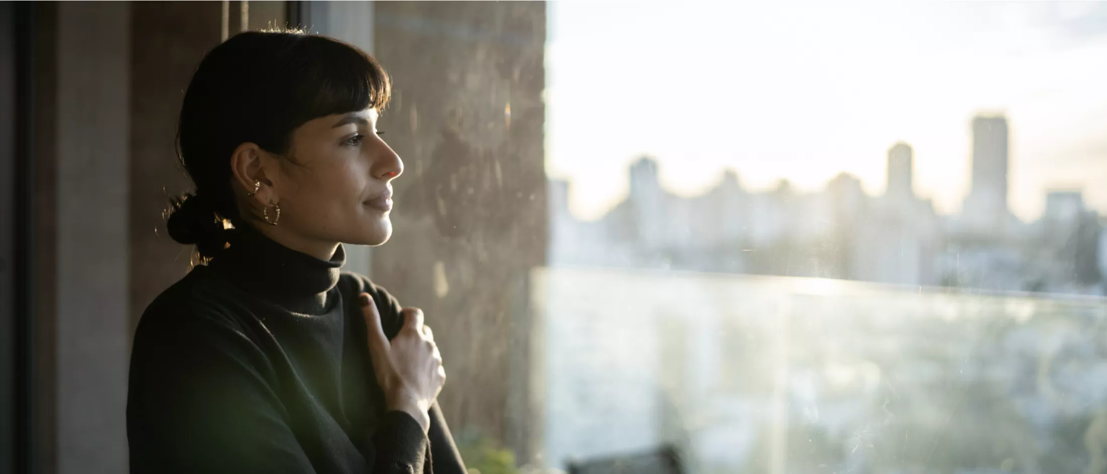 A woman wearing a dark sweater looks out a window as the sun comes up above a large city skyline.