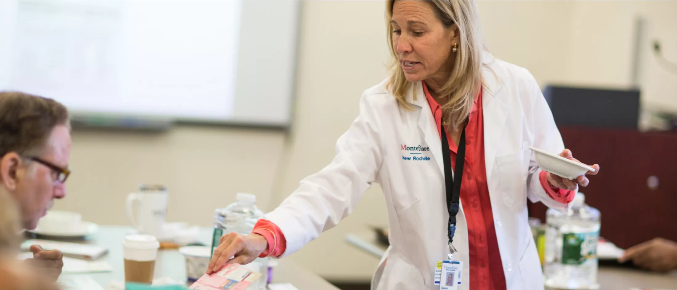 Doctor in lab coat holding a bowl in one hand, and in the other a cereal box that is being read by a seated man