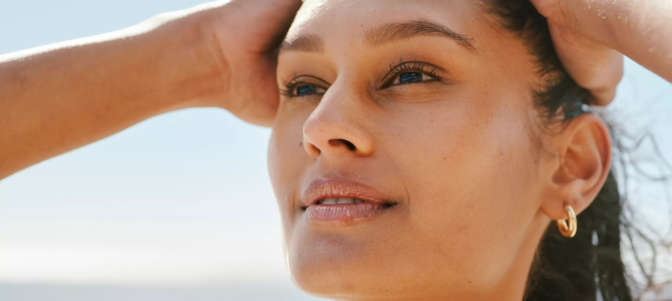 Confident woman with hands on head, gazing upward under sunlight, wearing gold hoop earring with a serene beach background.