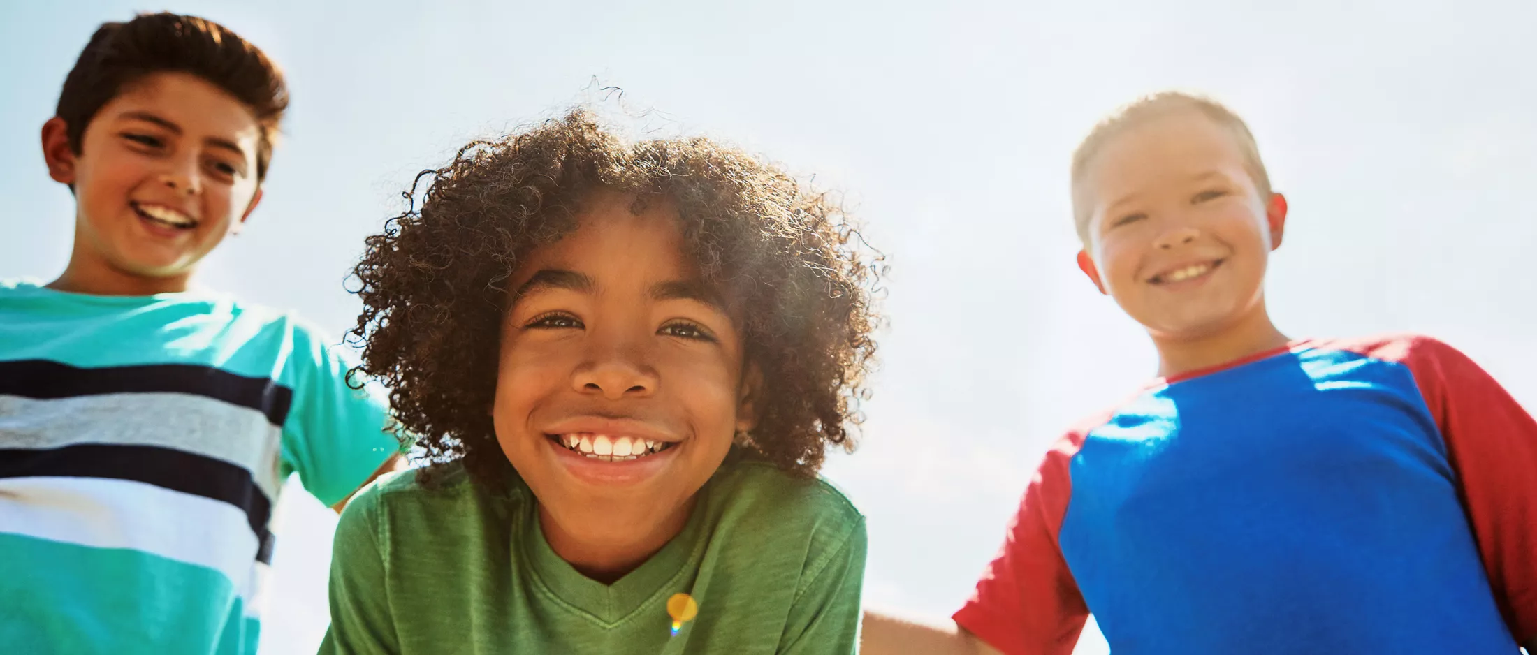 Three smiling children looking down at the camera under a bright blue sky