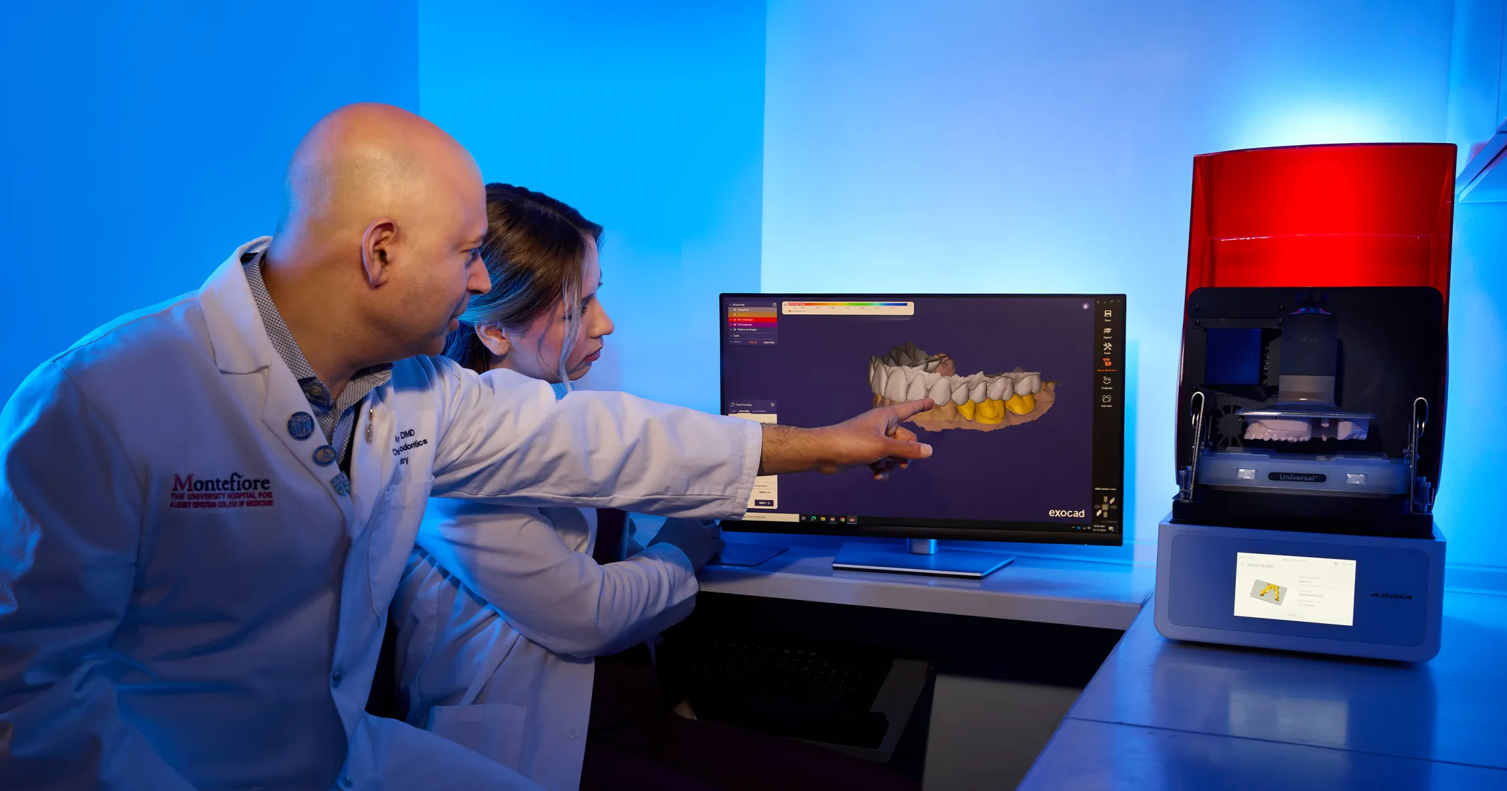 Two dental professionals reviewing a 3D model of teeth on a computer screen beside a 3D printer