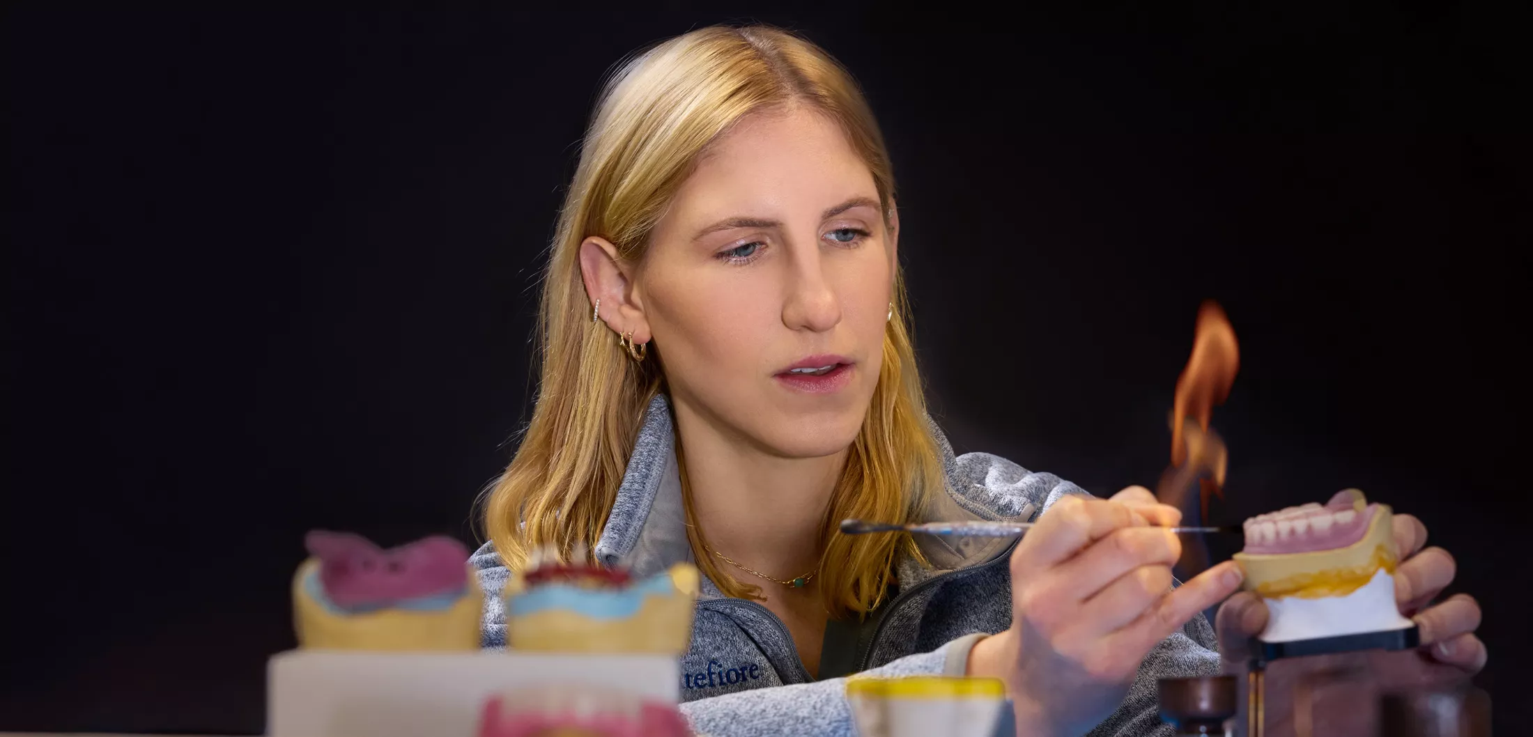 Woman carefully working on a dental mold using a metal tool near an open flame, with dental models on the table in front of her.