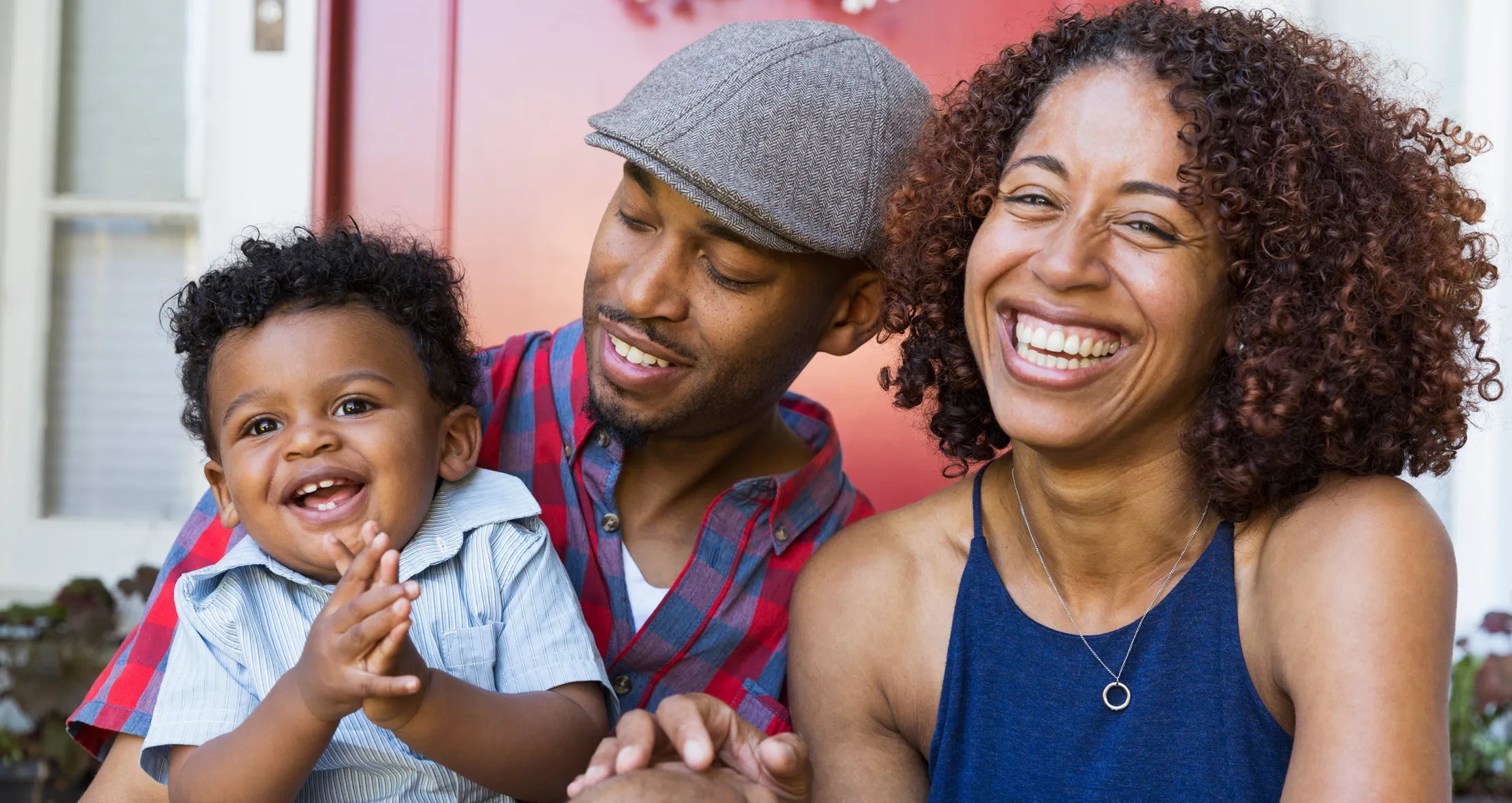 Smiling parents sit with their cheerful baby outside a red door.
