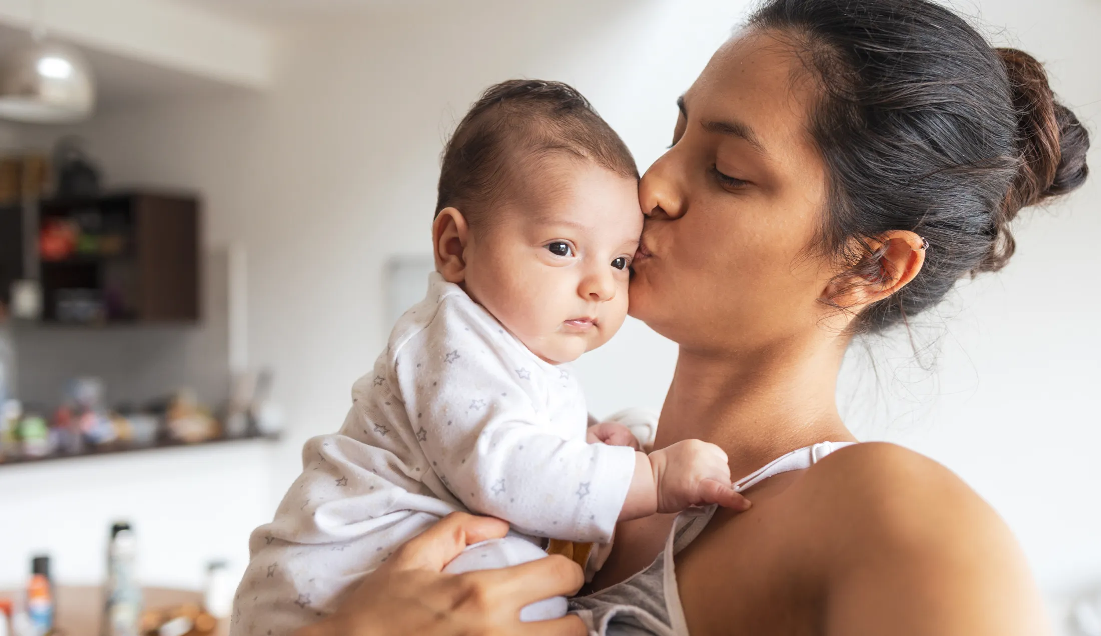 Woman kisses her baby on the cheek while holding the child in a cozy, home kitchen setting.