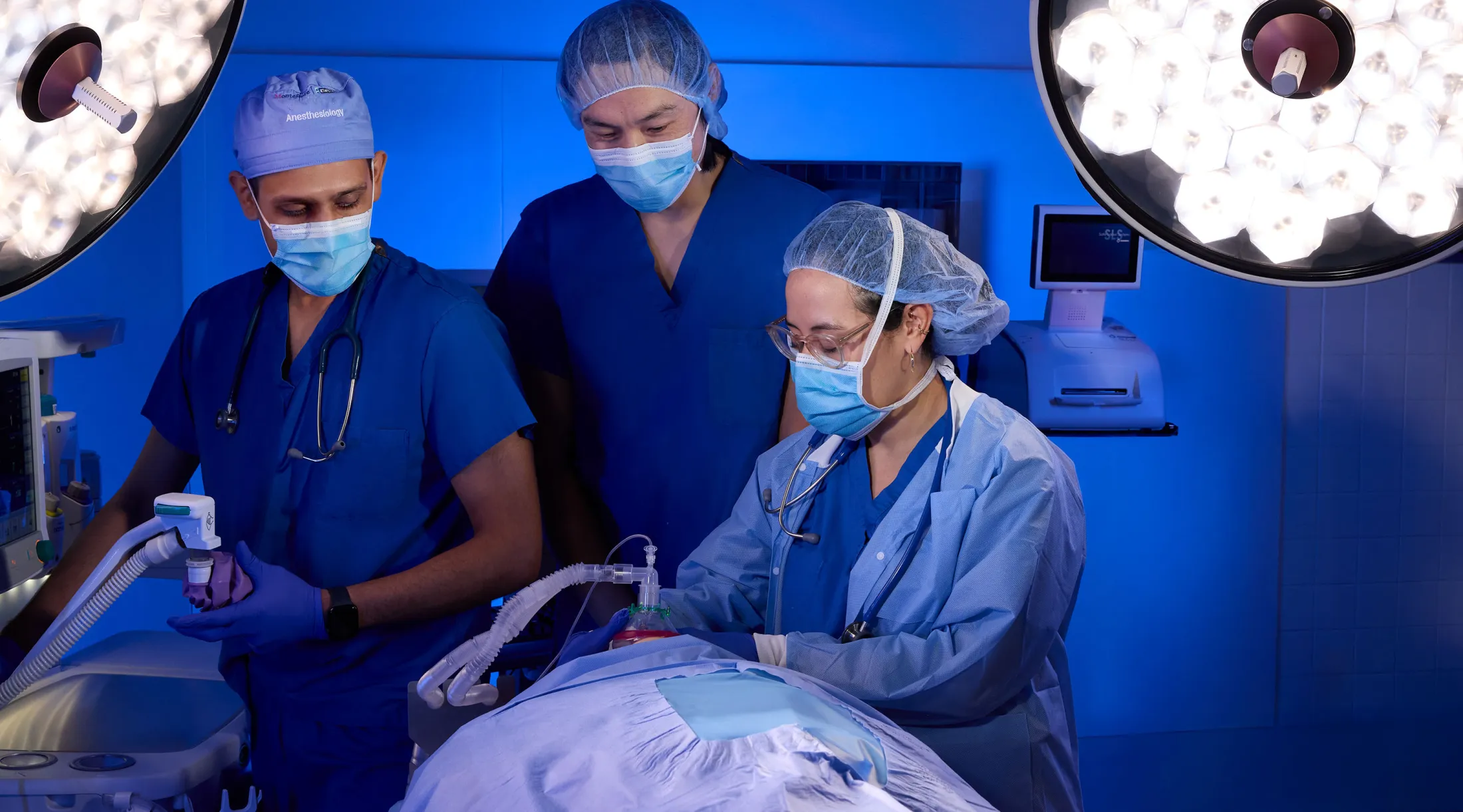  Three surgeons in blue scrubs and masks operate in a brightly lit hospital room.