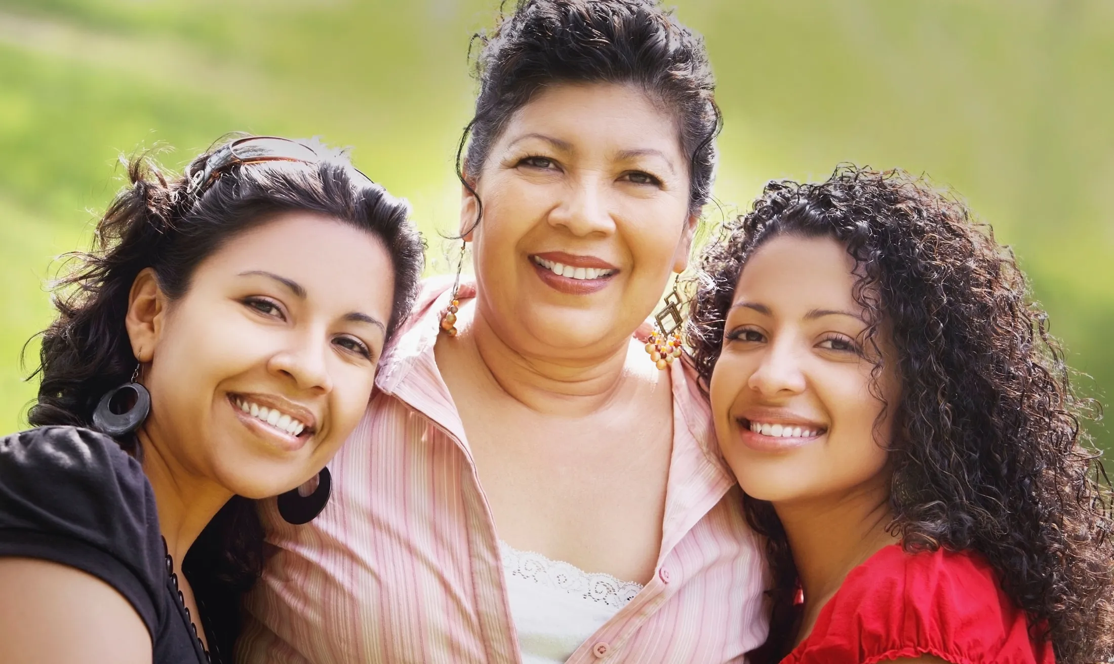 Three women smiling together outside on a sunny day, enjoying time together in a green park
