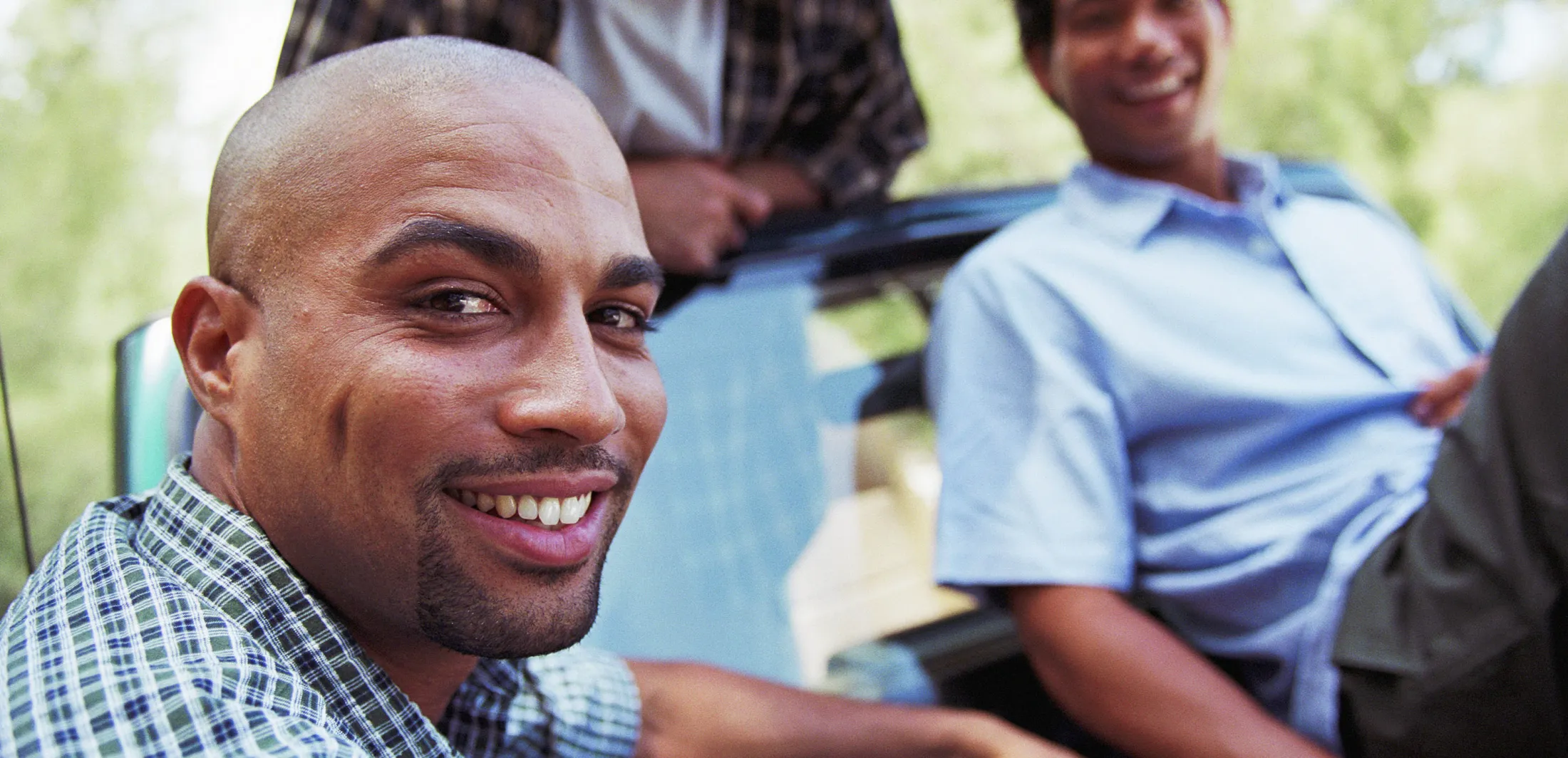 Smiling man sitting by a car with two friends in the background on a sunny day