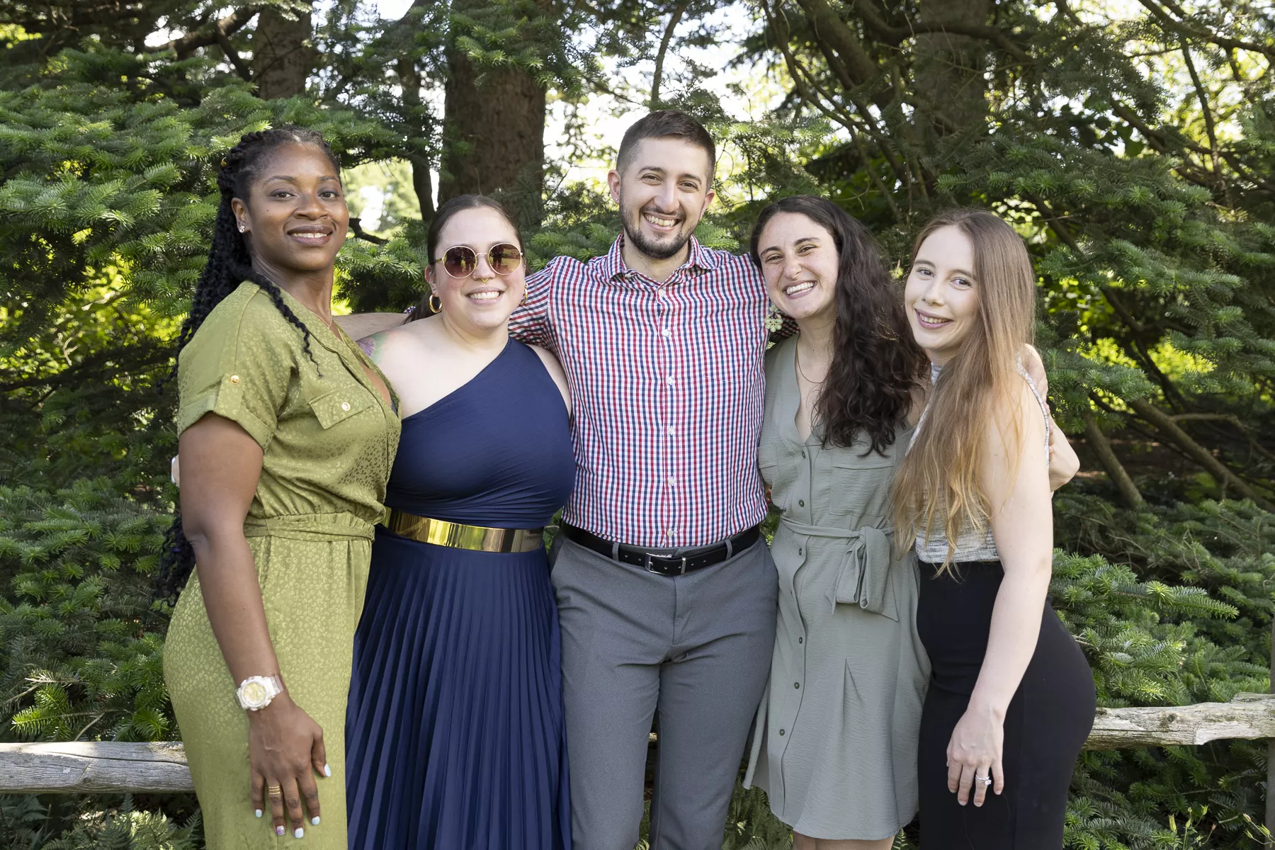 Five people posing outdoors, smiling in front of pine trees and dressed in colorful semi-formal attire.