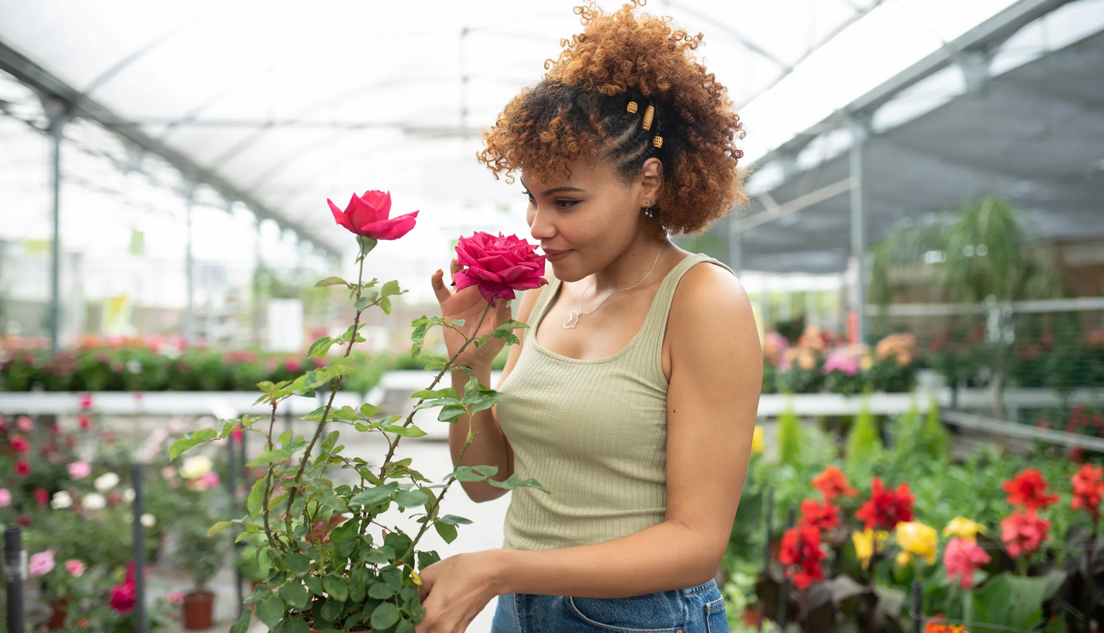 Woman smelling a pink rose in a greenhouse filled with blooming flowers.
