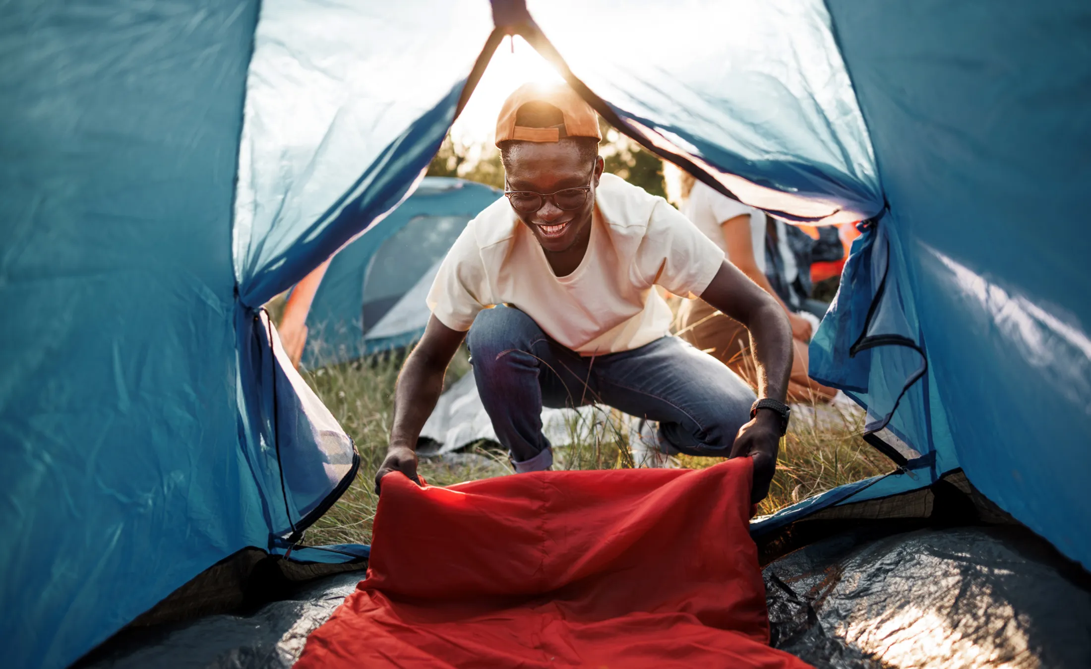 Man smiling while setting up a red sleeping bag inside a tent at a sunny campsite.