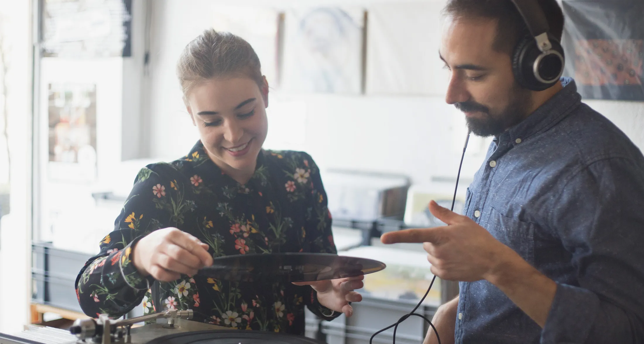 Woman smiling while placing a vinyl record on a turntable as a man wearing headphones watches.