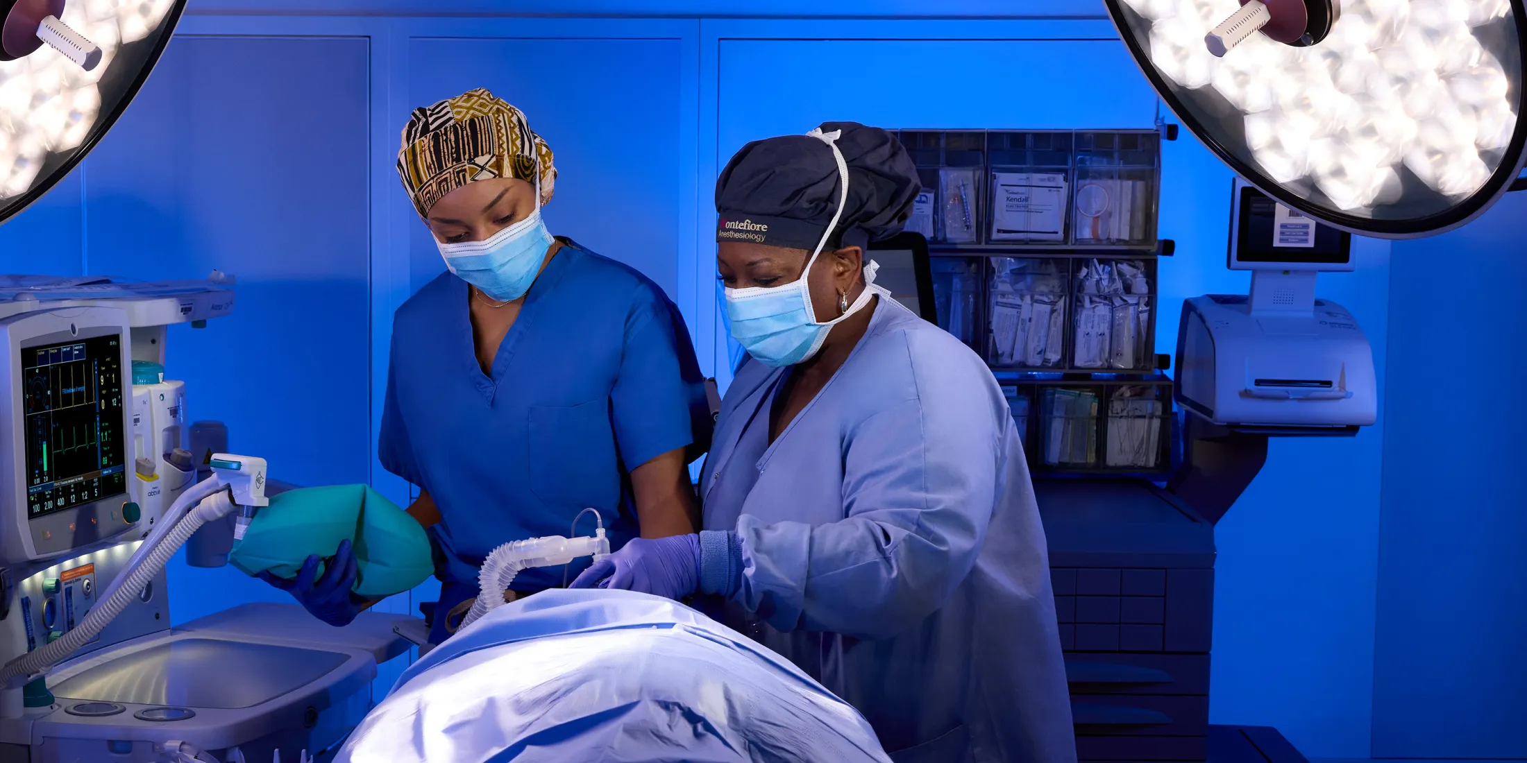 Two masked healthcare workers preparing a patient for surgery in an operating room under bright surgical lights