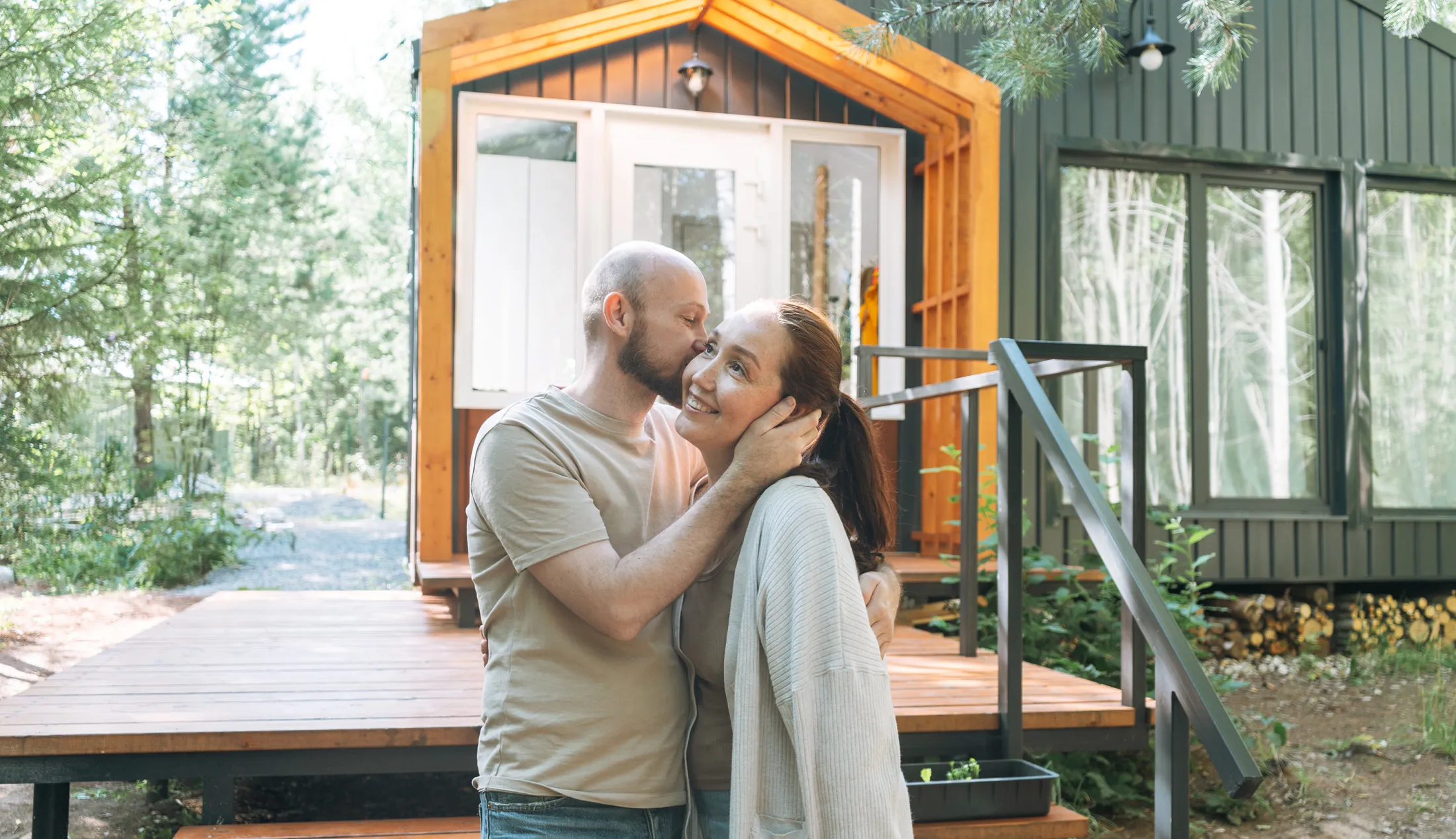 A man kisses a smiling woman on the cheek outside a cozy modern cabin surrounded by trees.