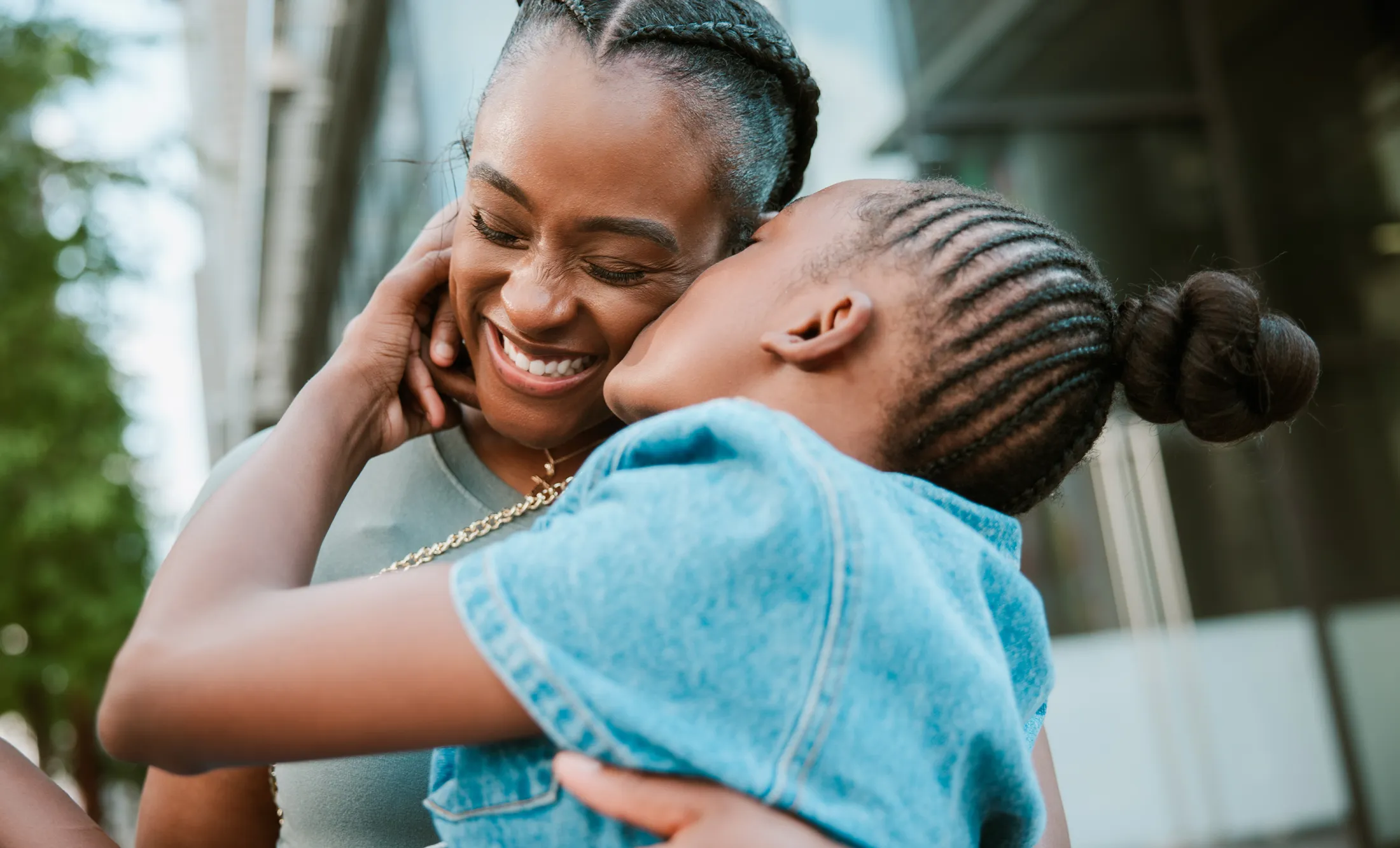 Close-up of a smiling woman receiving an affectionate hug and kiss on the cheek from a young girl outdoors.