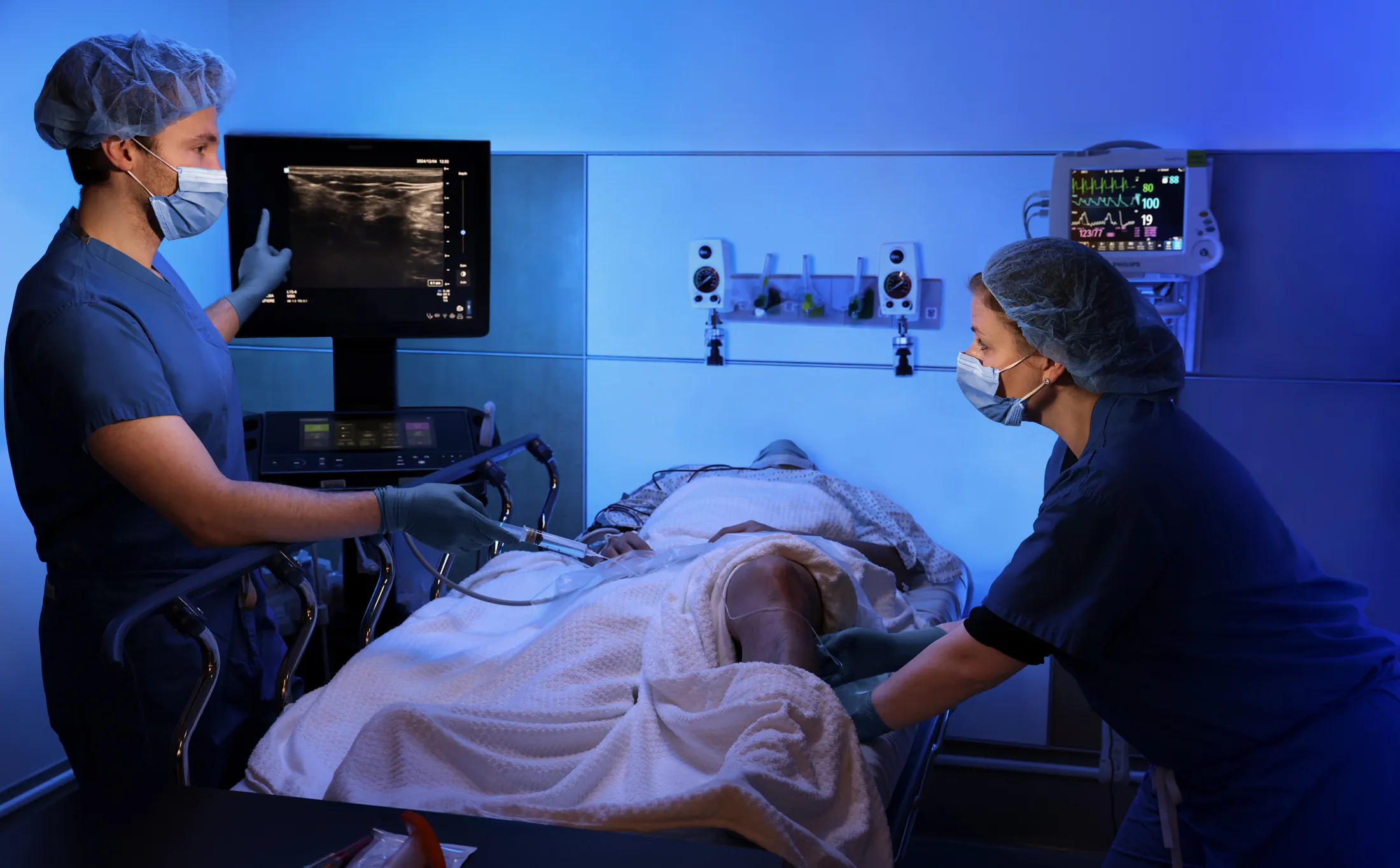Two medical professionals in surgical attire perform an ultrasound-guided procedure on a patient in a hospital room.