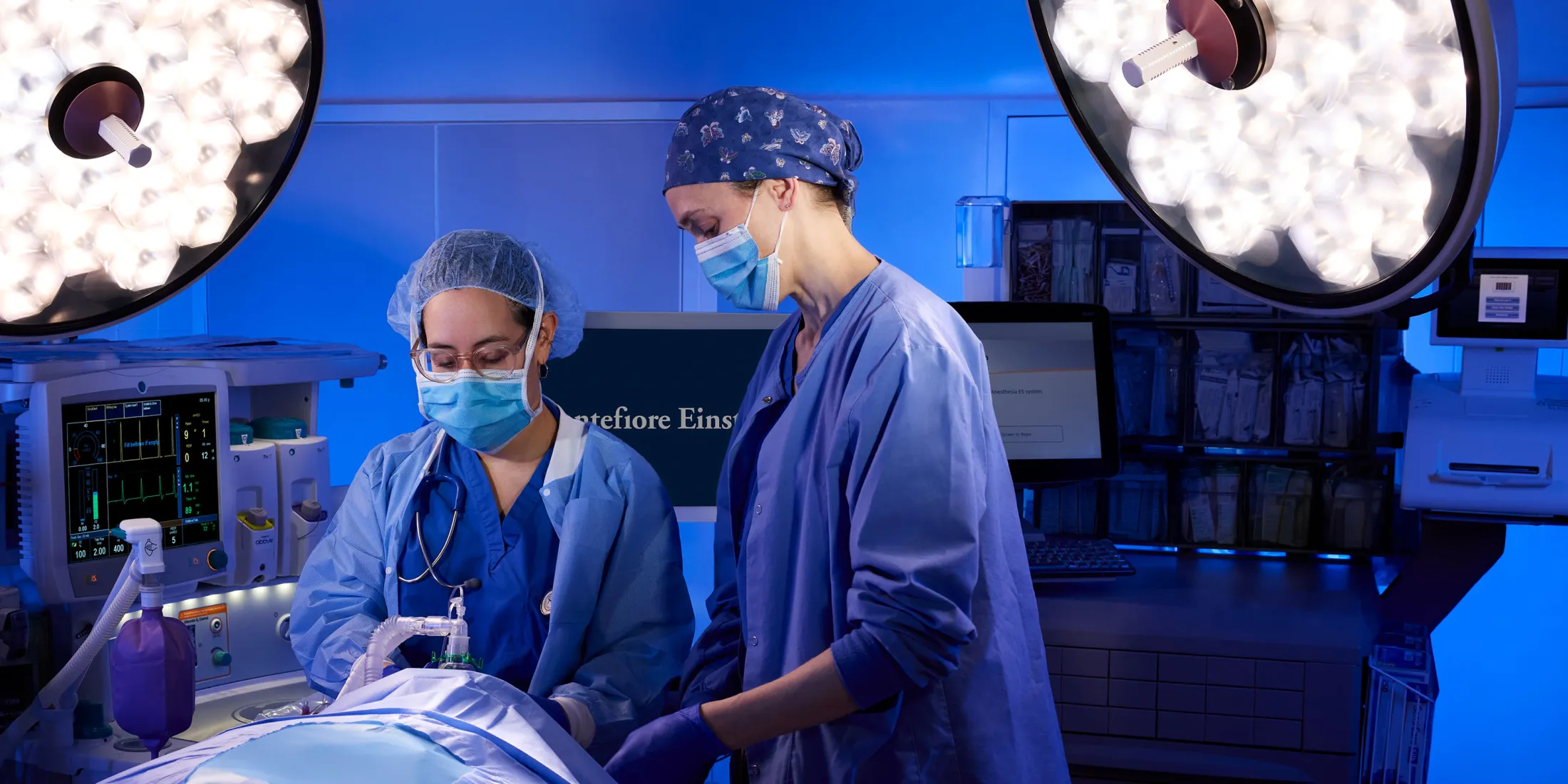 Two medical professionals in surgical attire work in a high-tech operating room under bright lights.