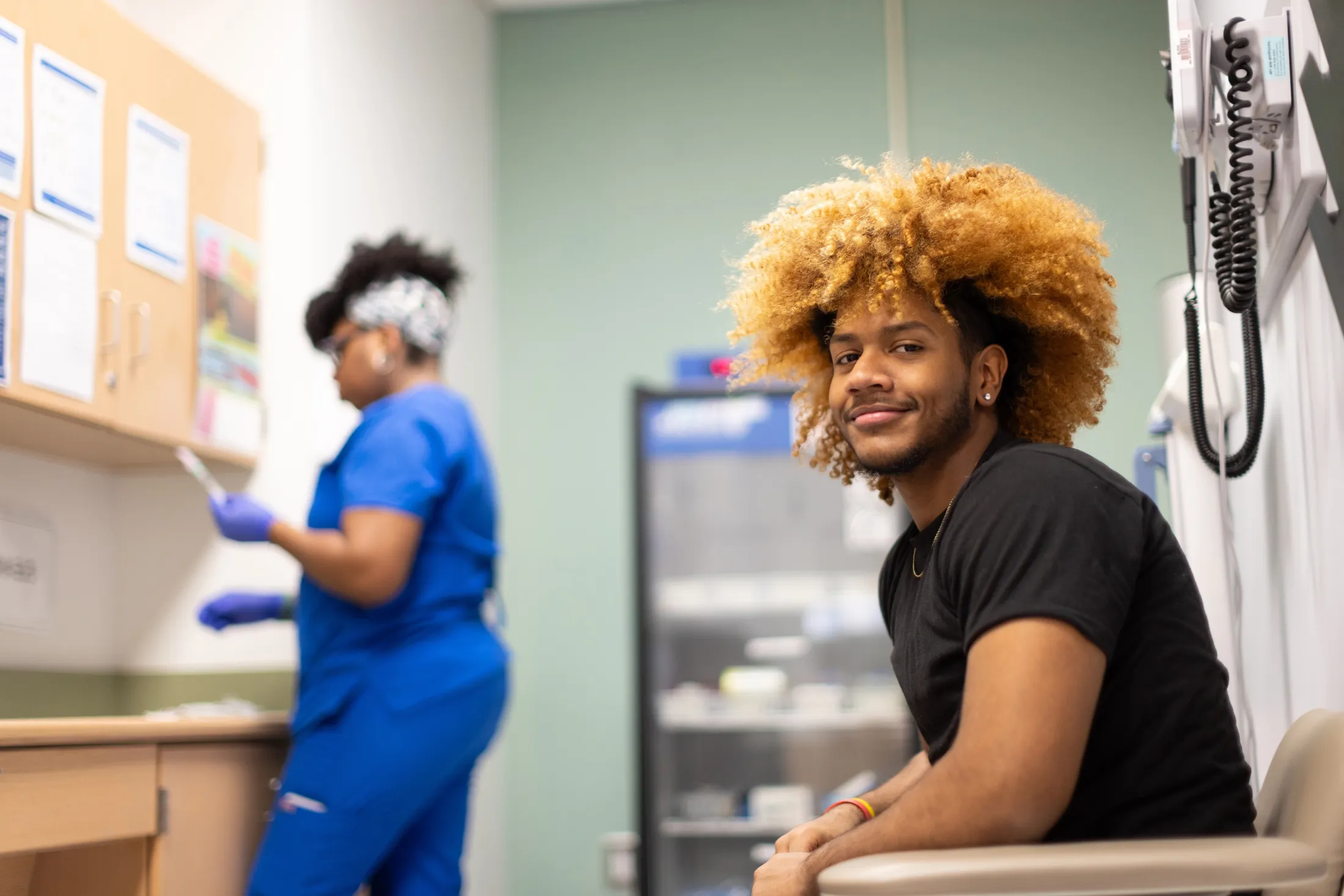 Young man with curly hair smiles while waiting in a medical setting, nurse working in background.