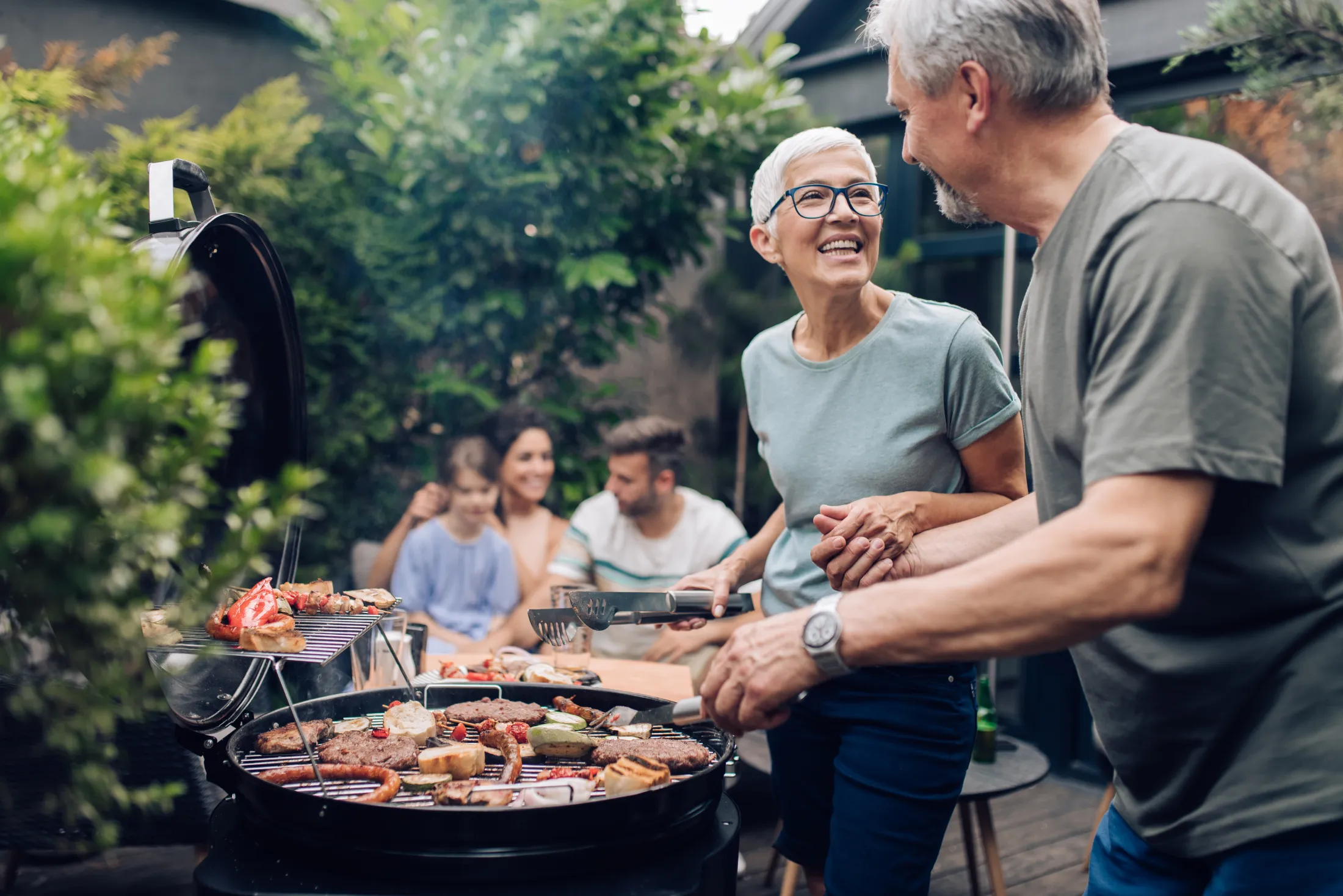 Older couple smiling and grilling food at an outdoor barbecue with family in the background.