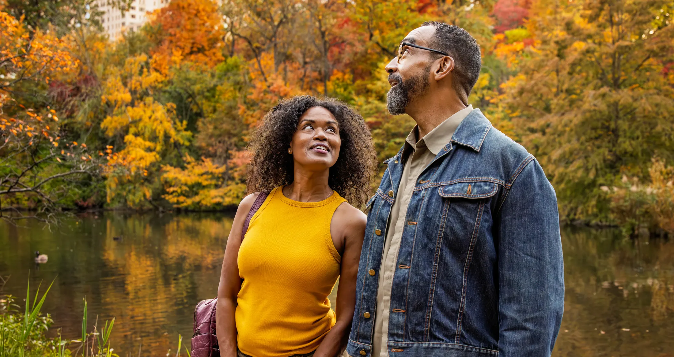 A middle-aged couple enjoys a walk by a lake surrounded by colorful autumn foliage in a park.