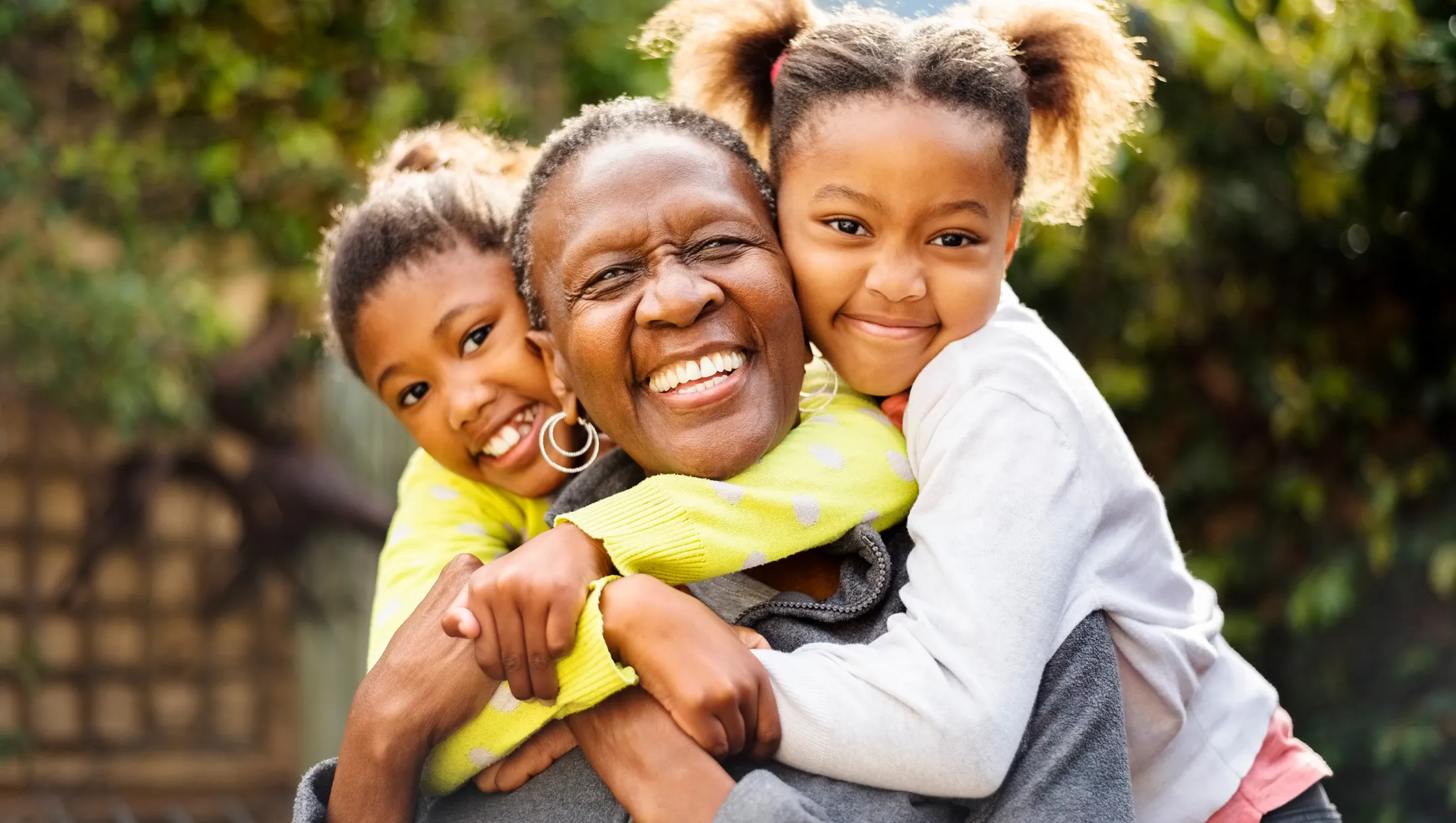 Joyful grandmother embracing two young granddaughters who are hugging her from both sides and smiling brightly.