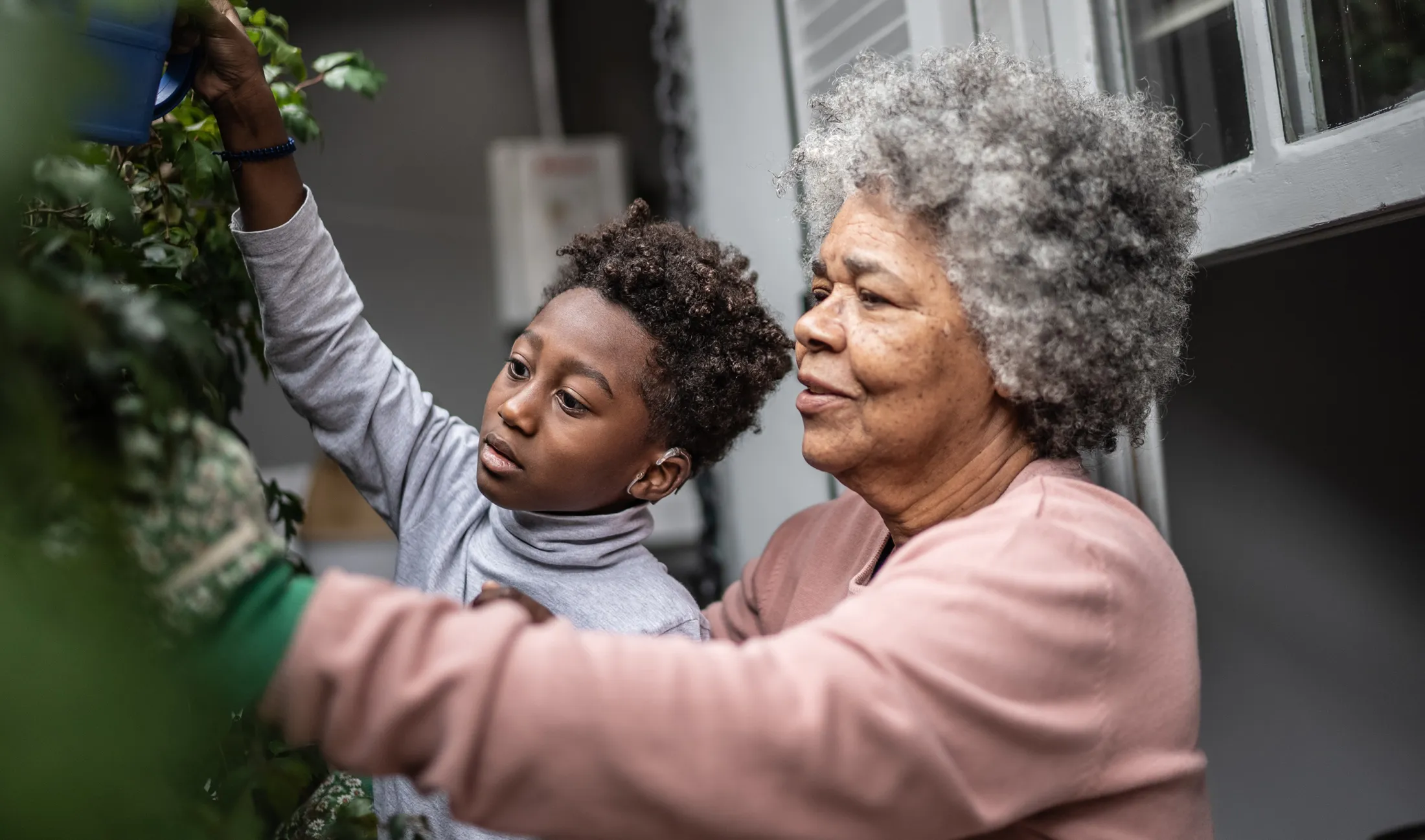 Elderly woman helping a young boy water plants in a garden, both focused on their task.