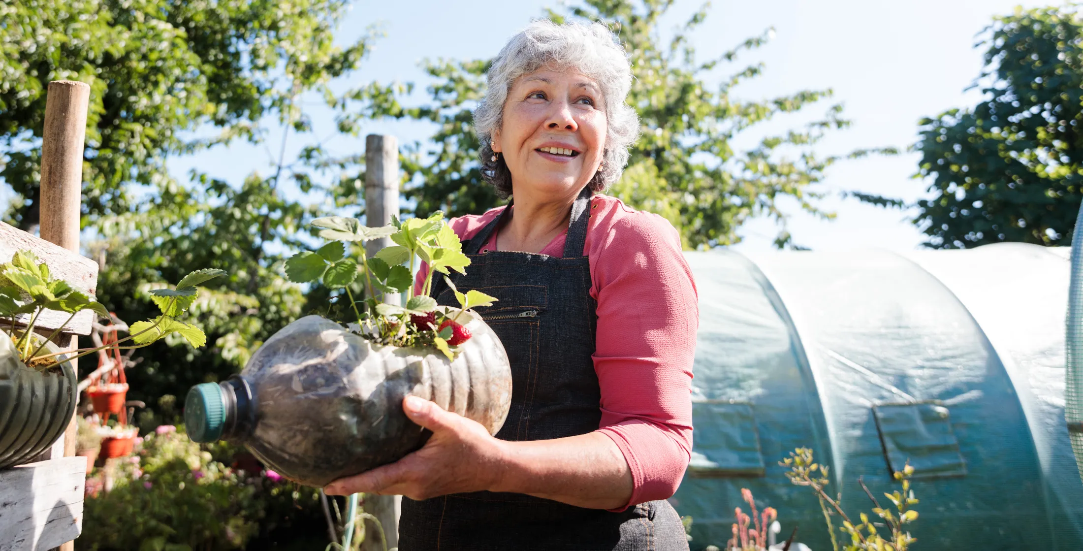 Older woman holding a handmade planter made from a plastic bottle in a lush garden, smiling under the sunlight.