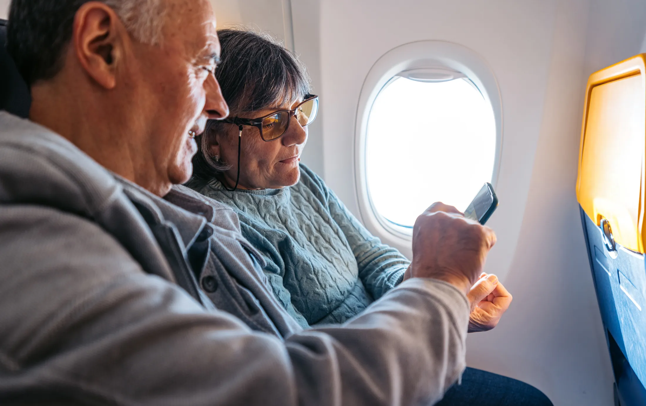 Older couple sitting side by side on an airplane, looking at a smartphone near the window.