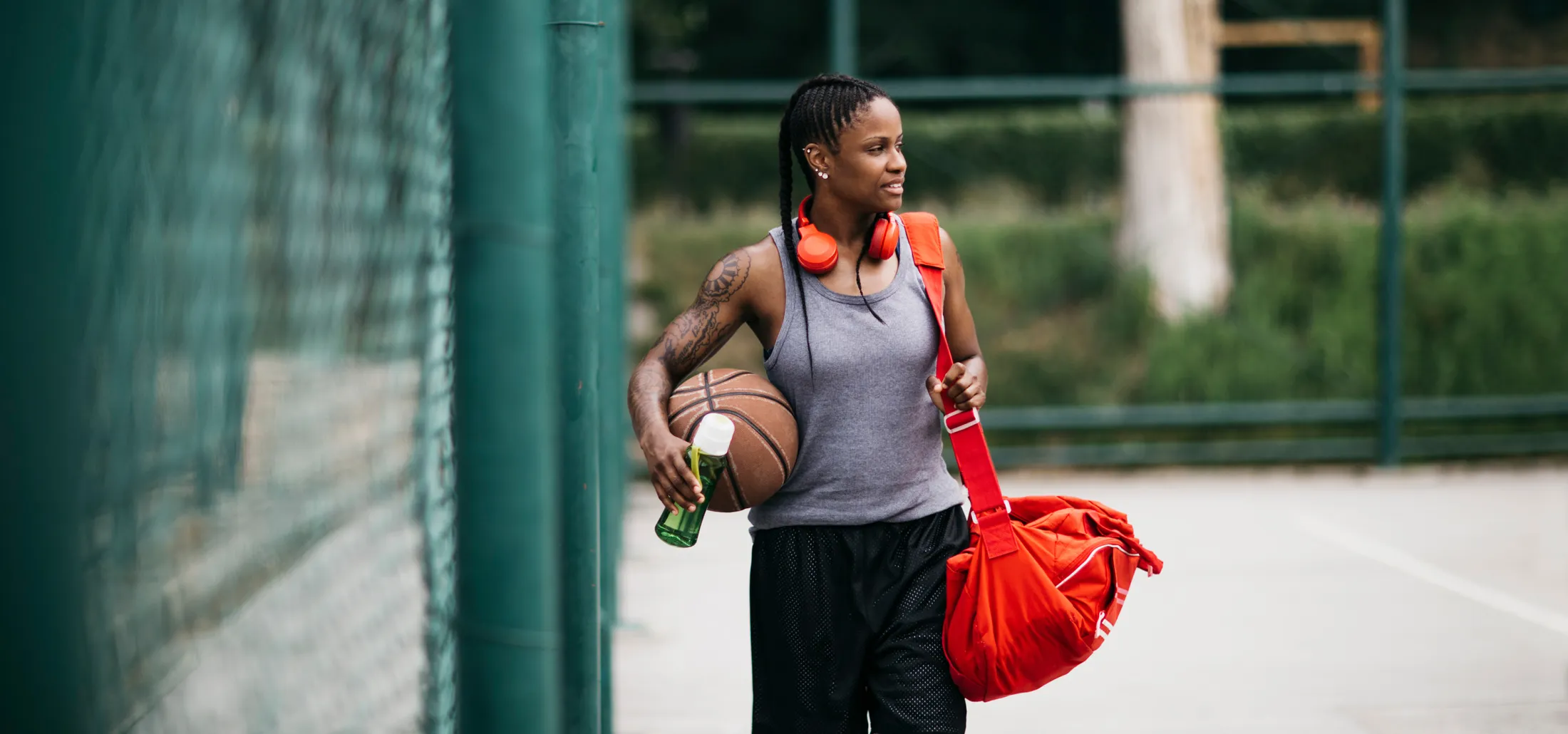 Woman walking on an outdoor basketball court carrying a basketball, a red duffel bag, and a drink bottle, with orange headphones around her neck.