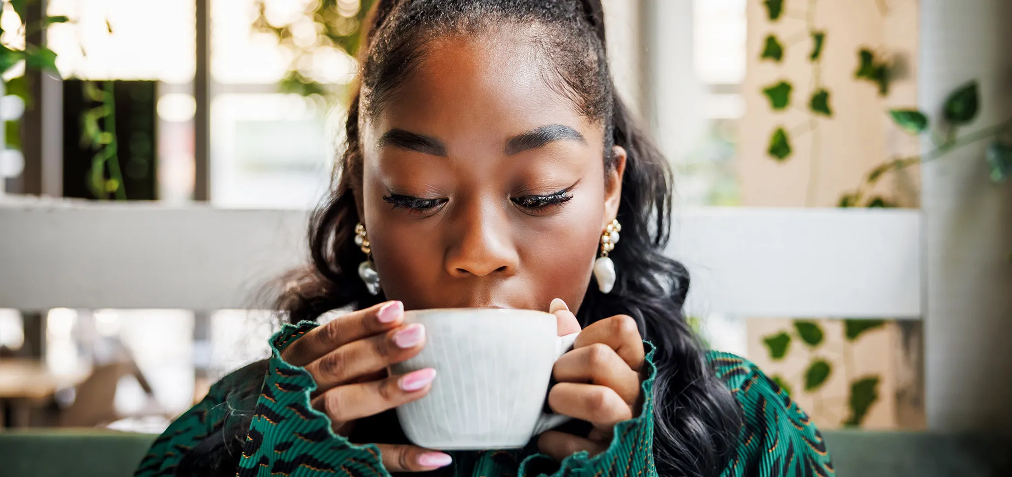 Woman with pearl earrings sipping from a white coffee mug in a cafe.
